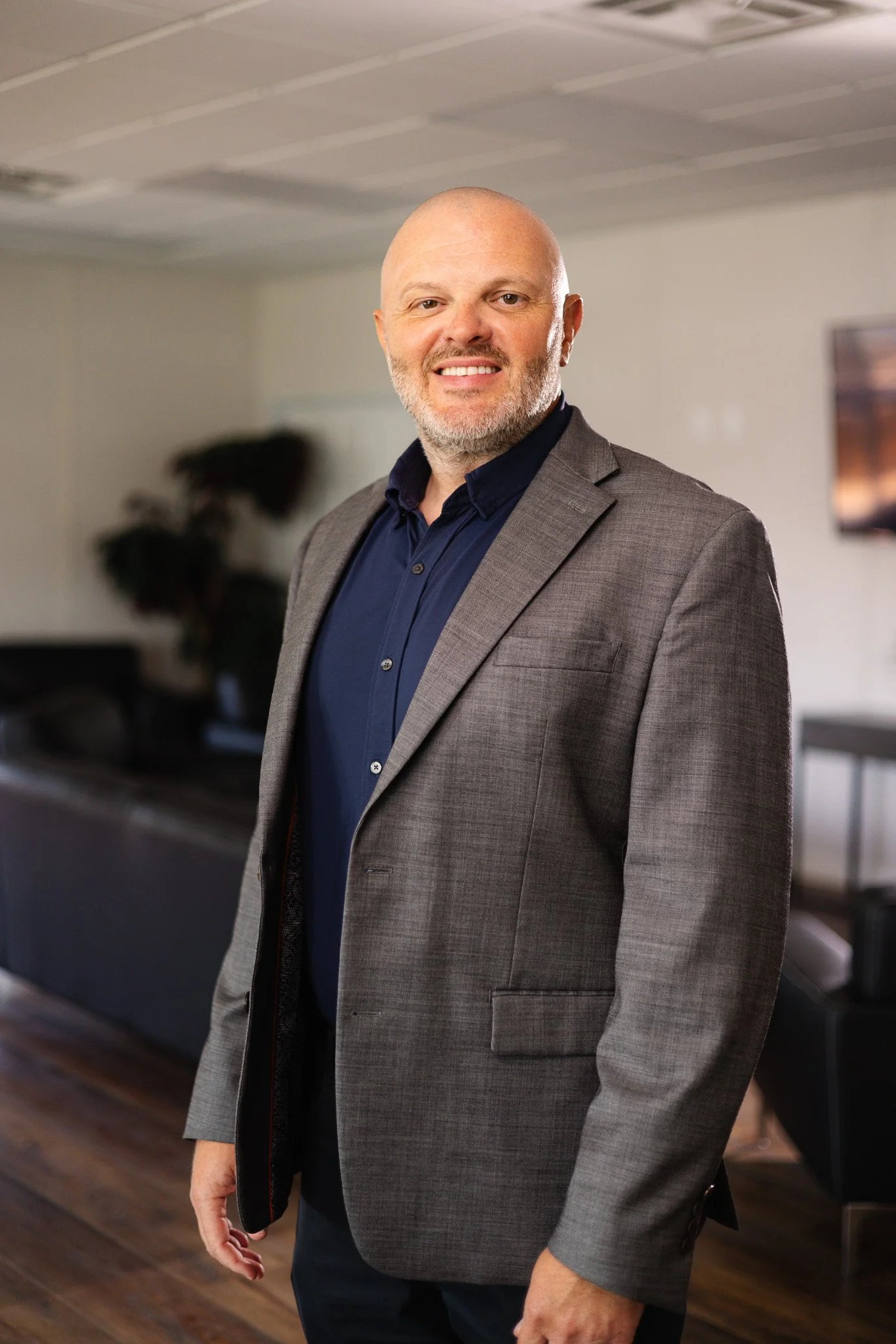 A man with a bald head, beard, and mustache wearing a dark plaid blazer and blue shirt, smiling with arms crossed against a plain gray background.