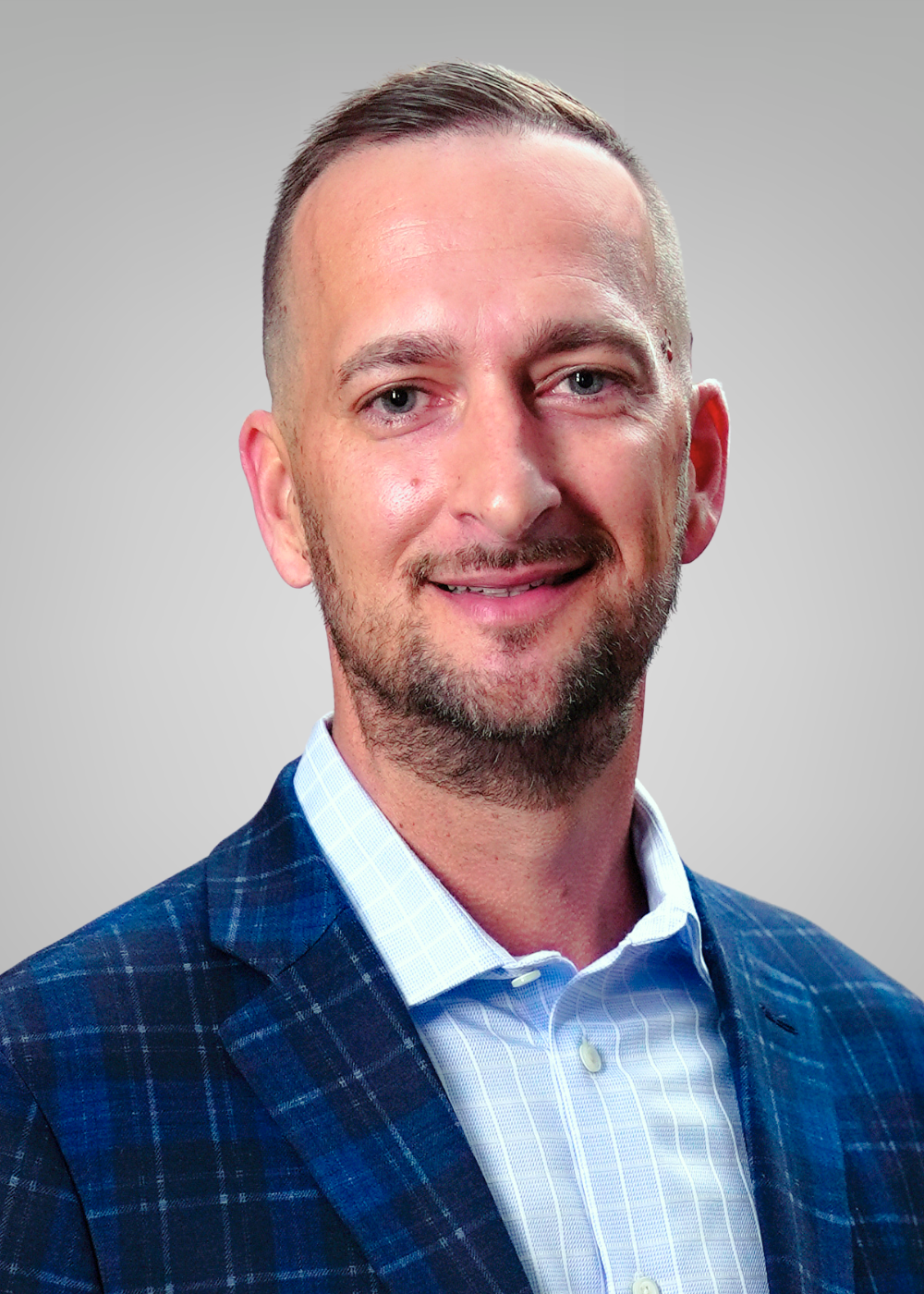 A headshot of a smiling man with short brown hair, a beard, and blue eyes wearing a white shirt and a blue checkered blazer against a light gray background.