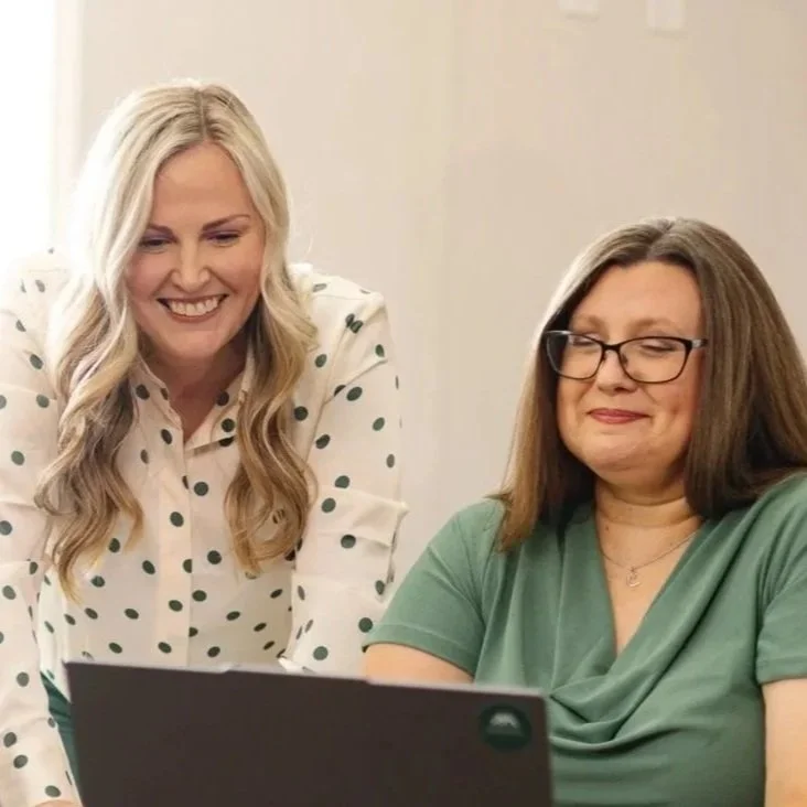 Two women looking at a laptop, one with blonde hair and wearing a white polka dot shirt, smiling, and the other with brown hair, glasses, and a green shirt, with a neutral expression.