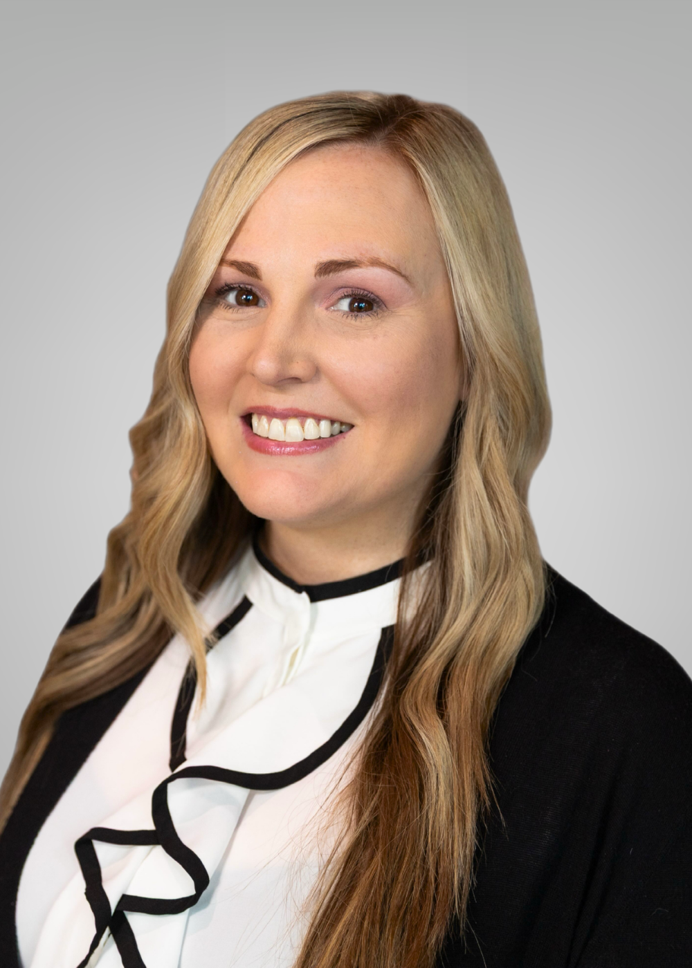 Headshot of a smiling woman with long, wavy blonde hair, wearing a black and white blouse with a black jacket, against a neutral gray background.