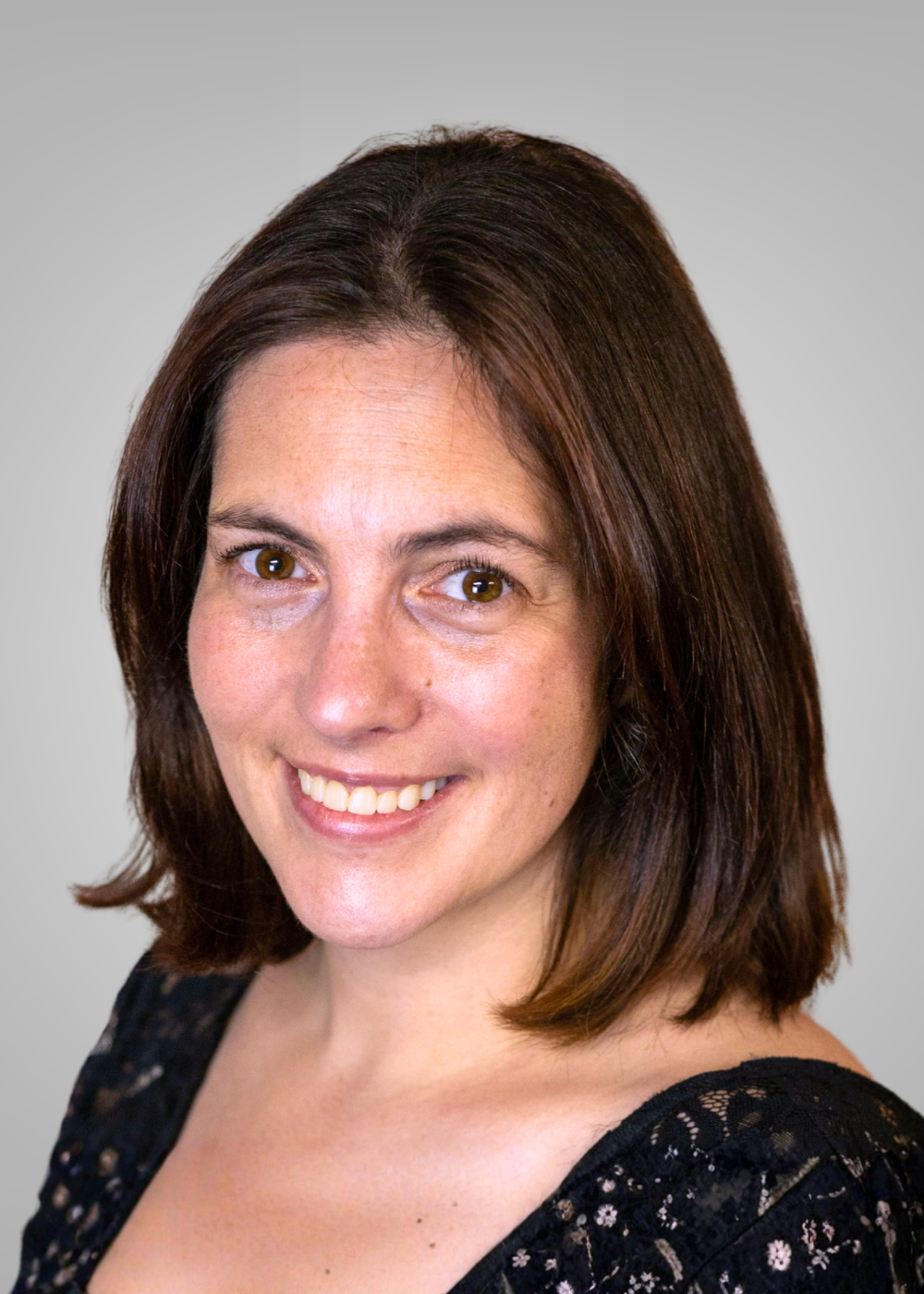A woman with shoulder-length dark brown hair, light skin, smiling, wearing a black lace top, against a plain light gray background.