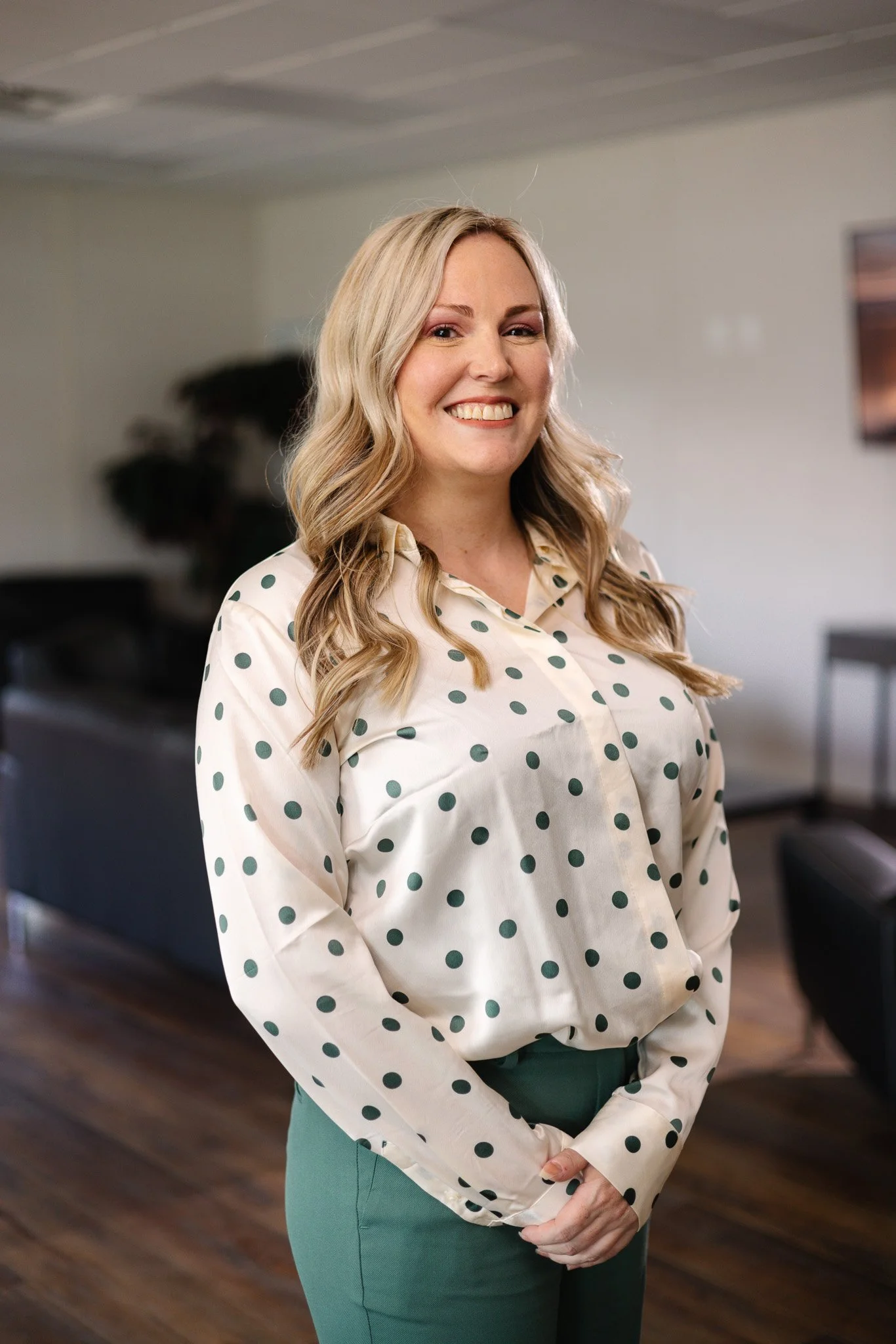 Headshot of a smiling woman with long, wavy blonde hair, wearing a black and white blouse with a black jacket, against a neutral gray background.