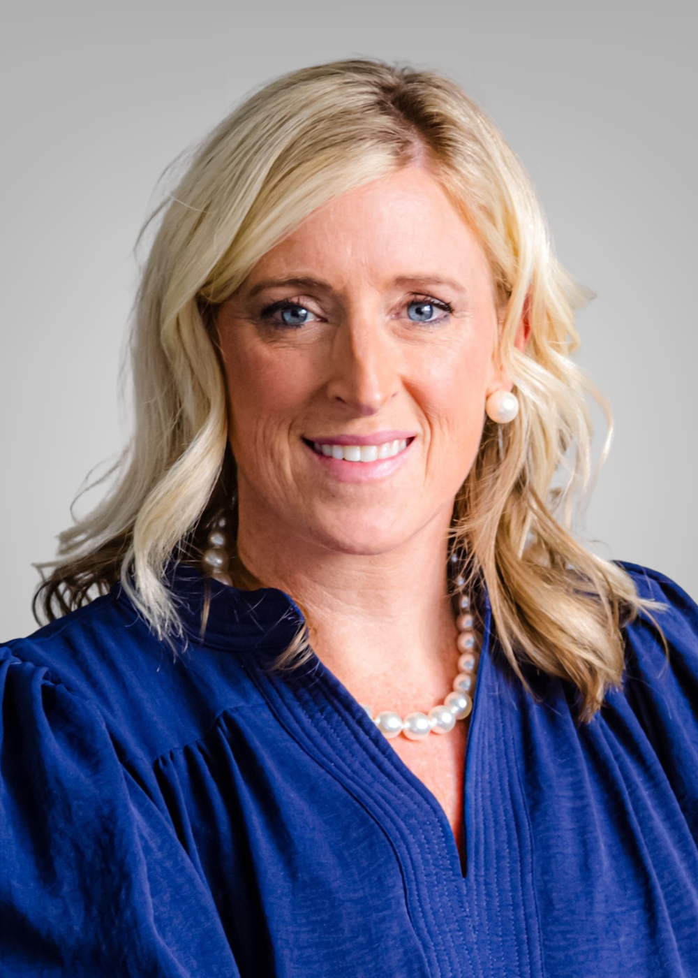 A smiling blonde woman wearing a navy blue blouse, pearl earrings, and a pearl necklace, against a light background.