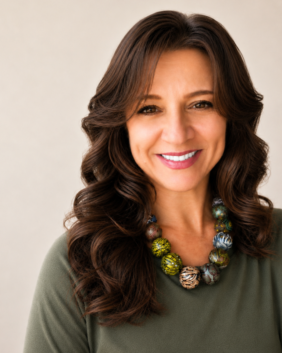 A woman with long, curly brown hair smiling at the camera, wearing a green top and a colorful beaded necklace. Lydia Spriggs - Founder Engage Enrich Empower