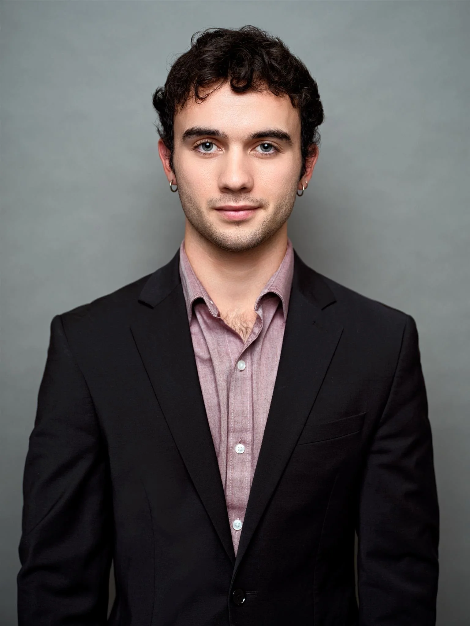 Portrait of a young man with curly brown hair wearing a black blazer over a pink shirt, against a gray background.