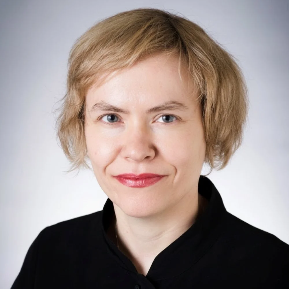 A woman with short blonde hair, fair skin, blue eyes, wearing a black top, smiling against a plain light gray background.