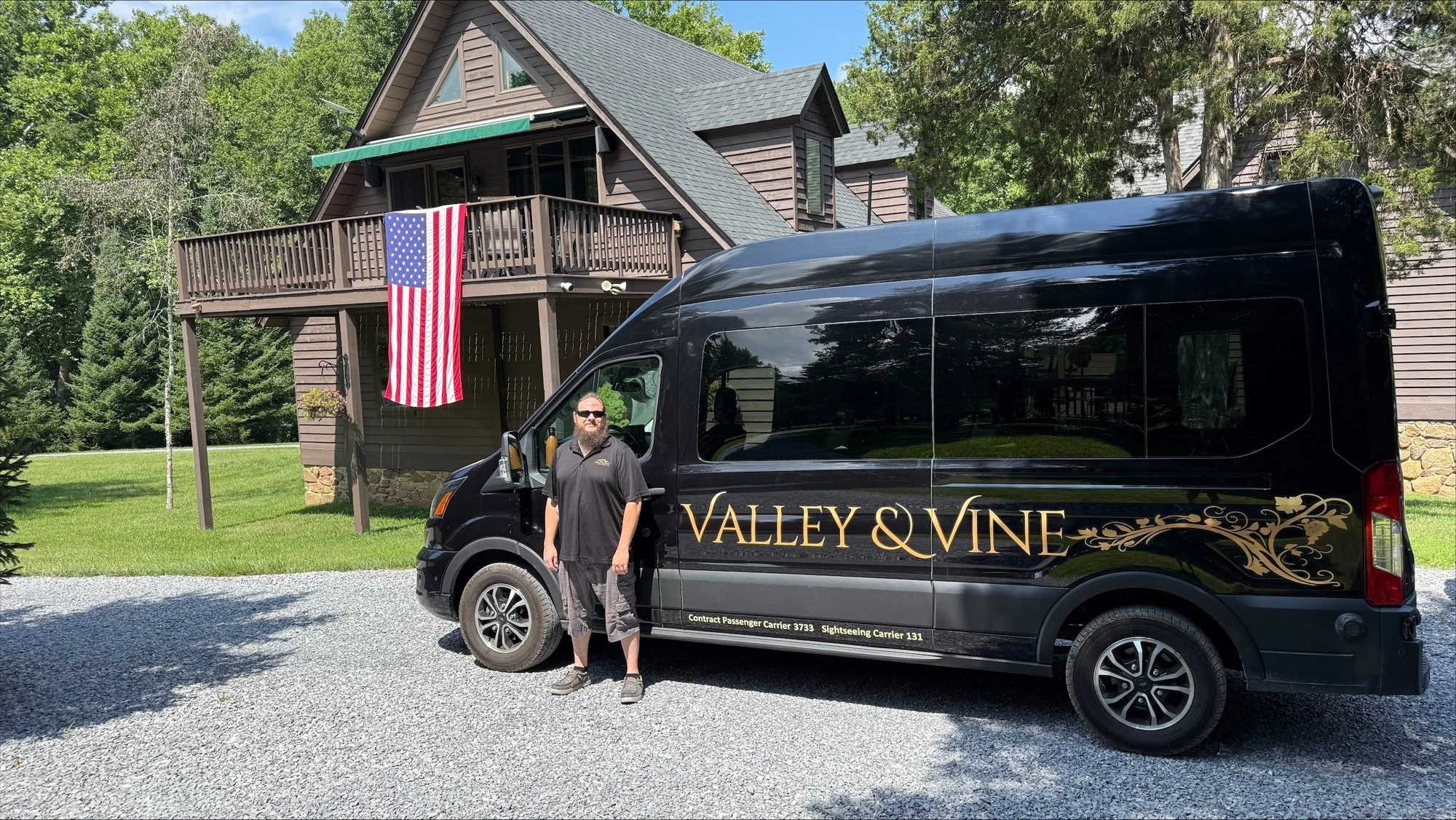 A man with a beard and sunglasses standing beside a black service van with 'Valley & Vine' written on the side, parked in front of a two-story house with a wooden exterior, a balcony, and an American flag hanging from a flagpole.
