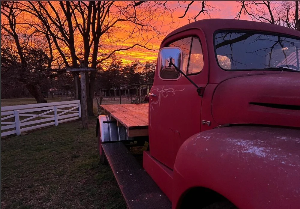 Old red truck with a wooden bed parked on a grassy area with trees and a white fence, during a colorful sunset sky.