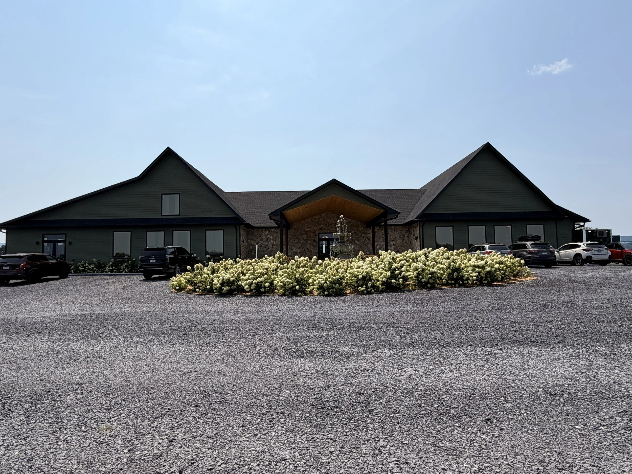 A large building with a dark green exterior, multiple windows, and a stone facade at the entrance, surrounded by a parking lot filled with cars and a flower bed with white flowers in the foreground. The building has a peaked roof and a small covered porch at the entrance. The sky is clear and blue.