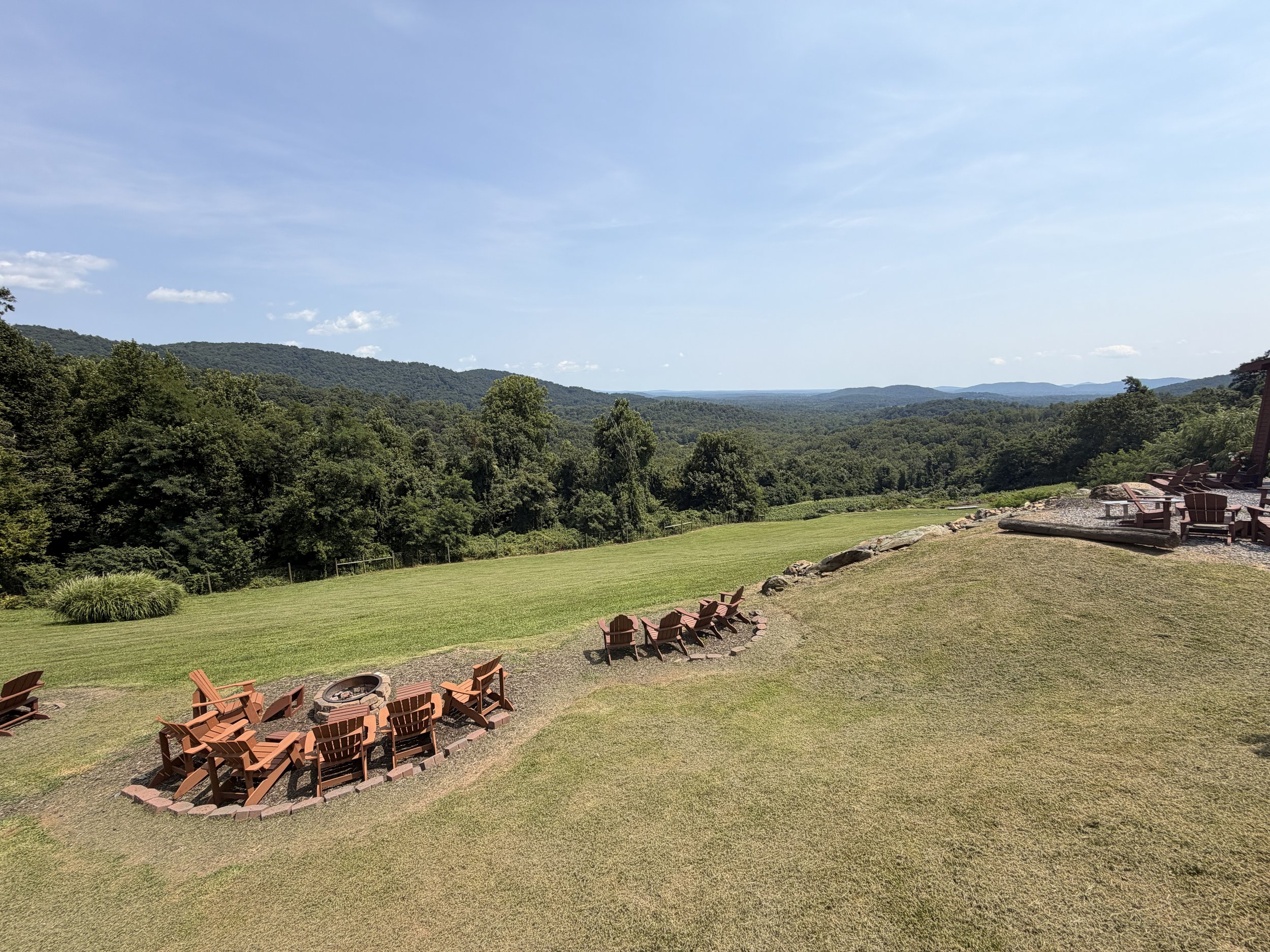 A scenic outdoor landscape with a grassy area, a fire pit with chairs around it, and a view of distant hills and trees under a blue sky.