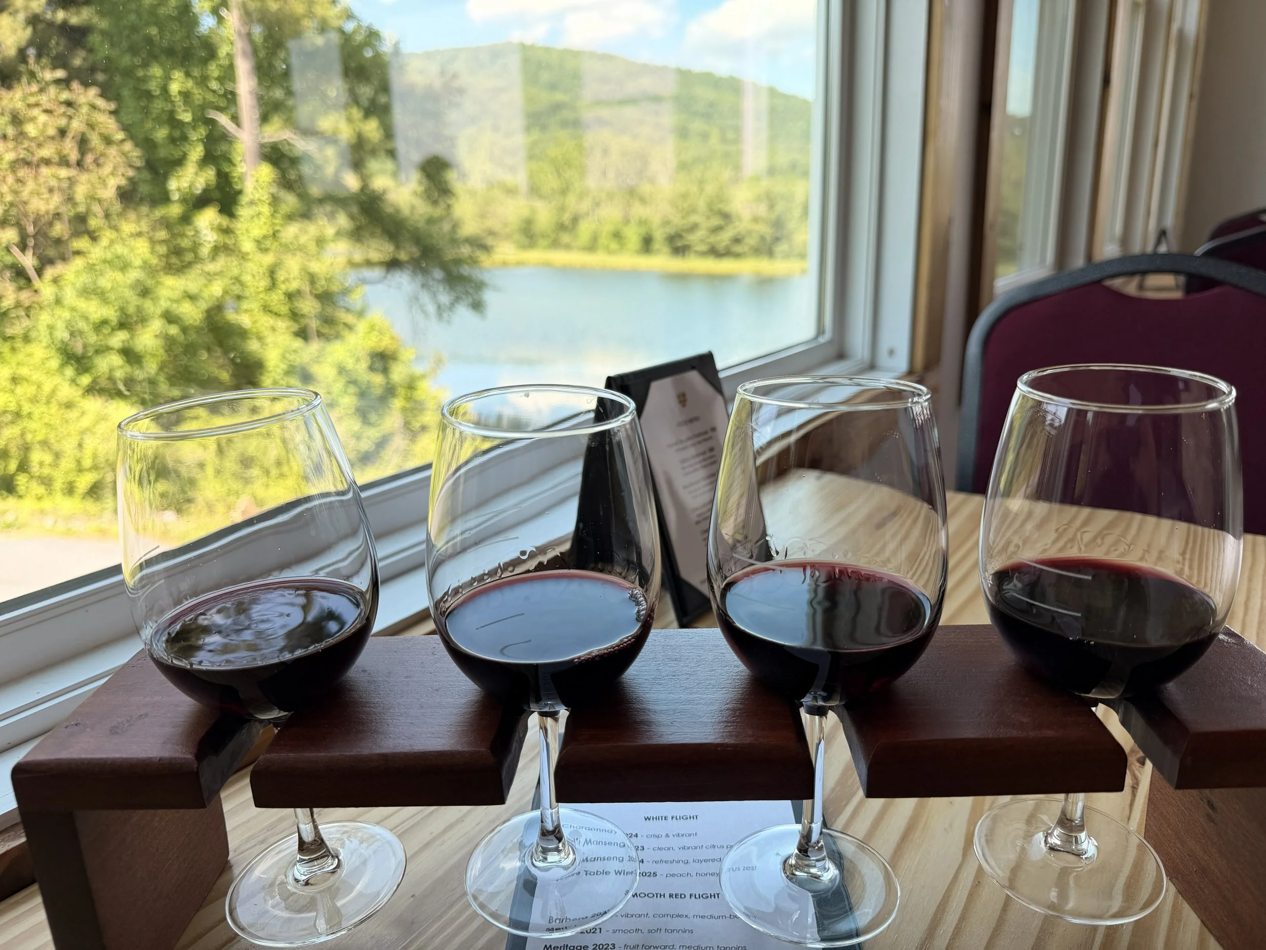 Four glasses of red wine on a wooden table with a view of a lake and green trees through a large window in the background.