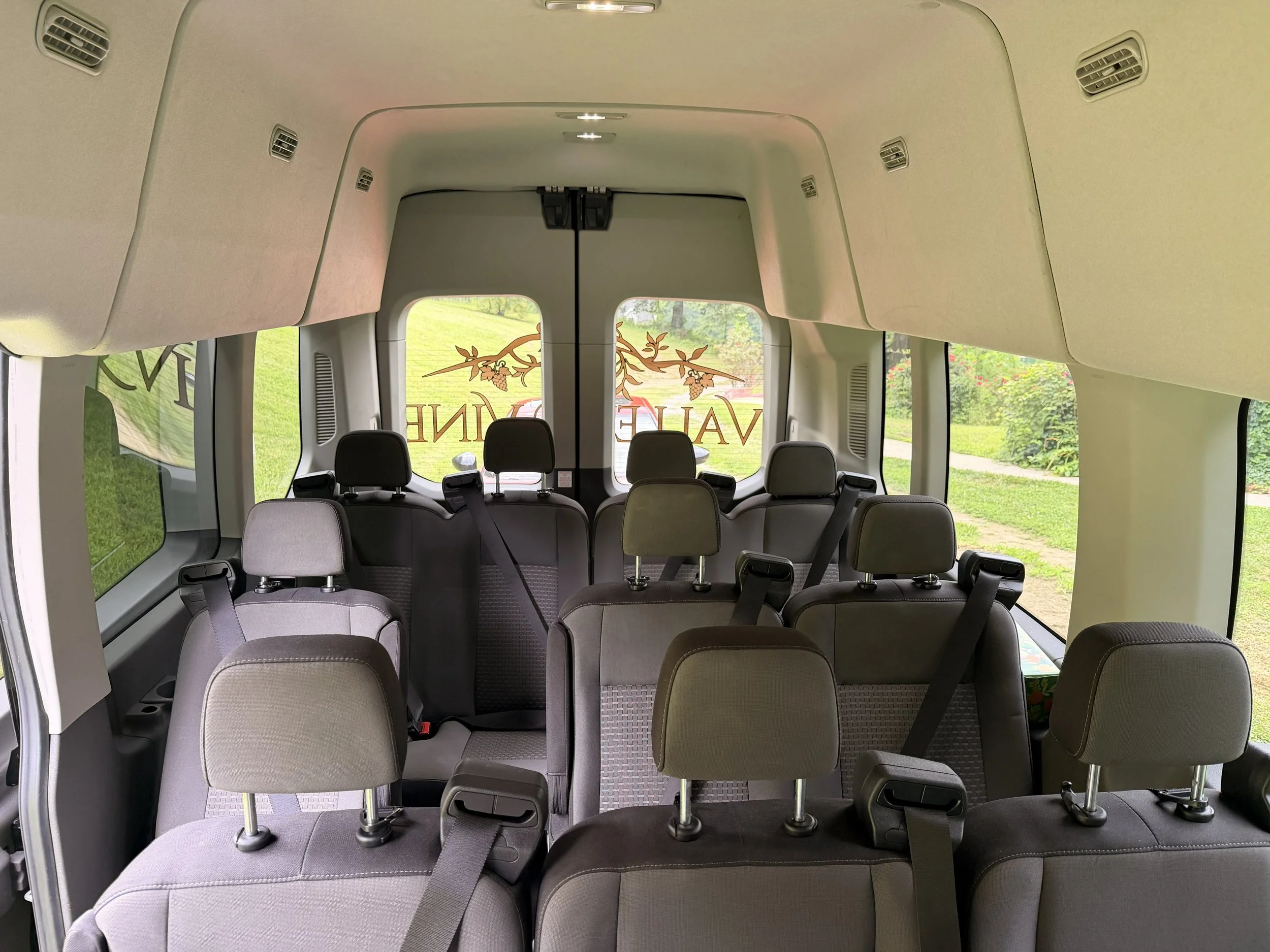 The interior of a passenger van with several rows of gray upholstered seats, black headrests, and seatbelts. The back of the van has large windows with a decorative tree and vine graphic on the outside. The ceiling has air vents and lights.