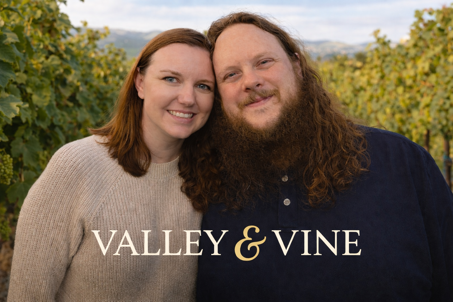 A smiling couple with the woman having shoulder-length red hair and the man with long curly red hair and a beard, standing in a vineyard with green grapevines and hills in the background, overlaid with the text "Valley & Vine."
