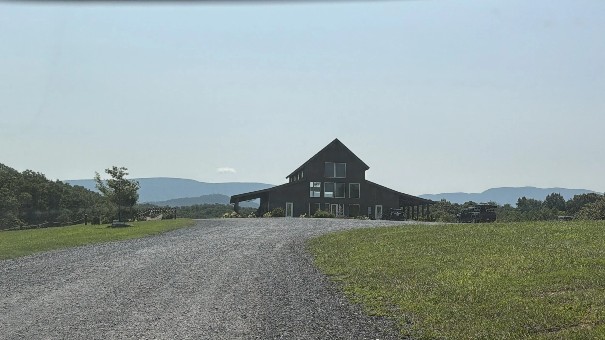 A rural scene with a gravel driveway leading to a modern black house with large windows, surrounded by green grass and trees, with mountains in the background on a clear day.