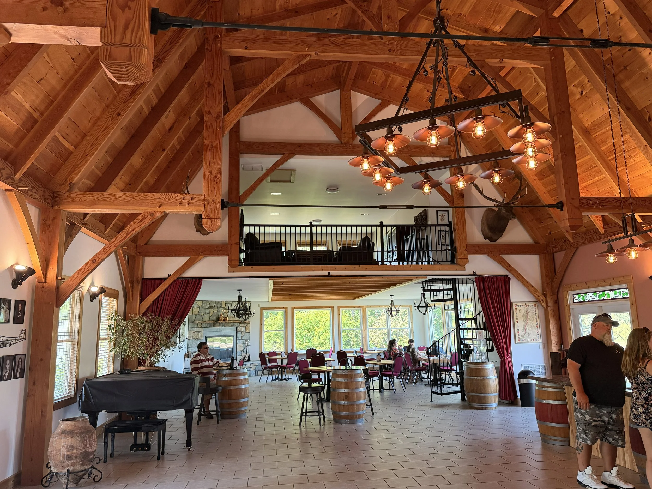 Interior of a rustic lodge with wooden beams and large windows, featuring a hanging chandelier with multiple Edison bulbs, people sitting and standing, and a fireplace in the background.