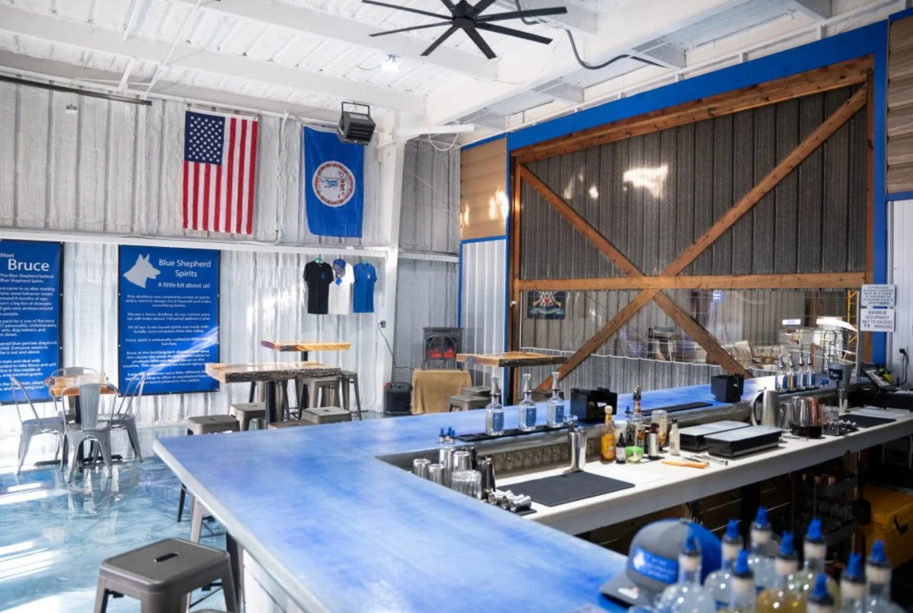 Interior of a bar or brewery with an open counter, bar stools, and a brewing tank behind the counter. Decorations include American and Missouri flags, and blue shepherd spirit signs and T-shirts on the wall.