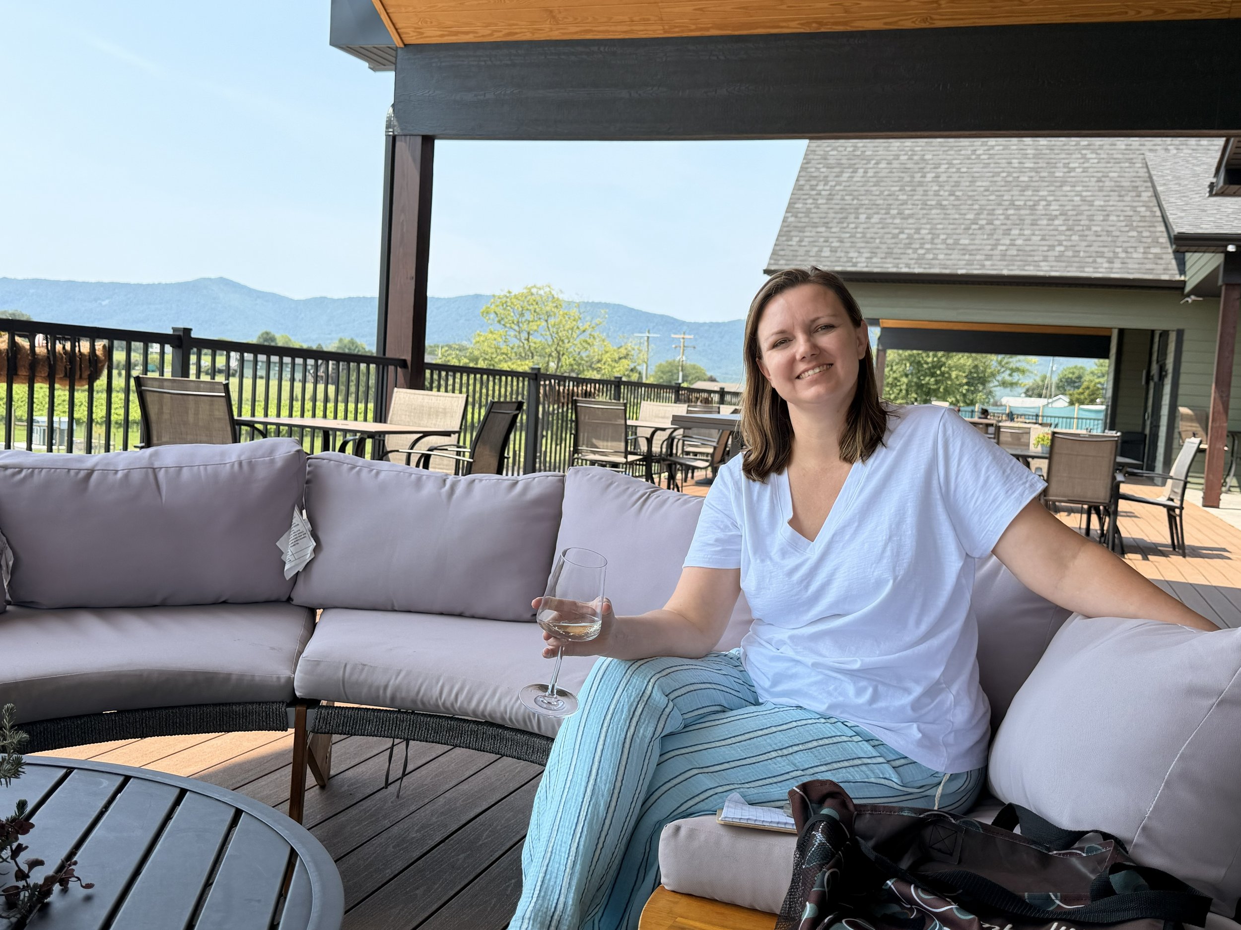 A woman smiling and sitting on a outdoor patio couch holding a glass of white wine with a mountain view in the background.