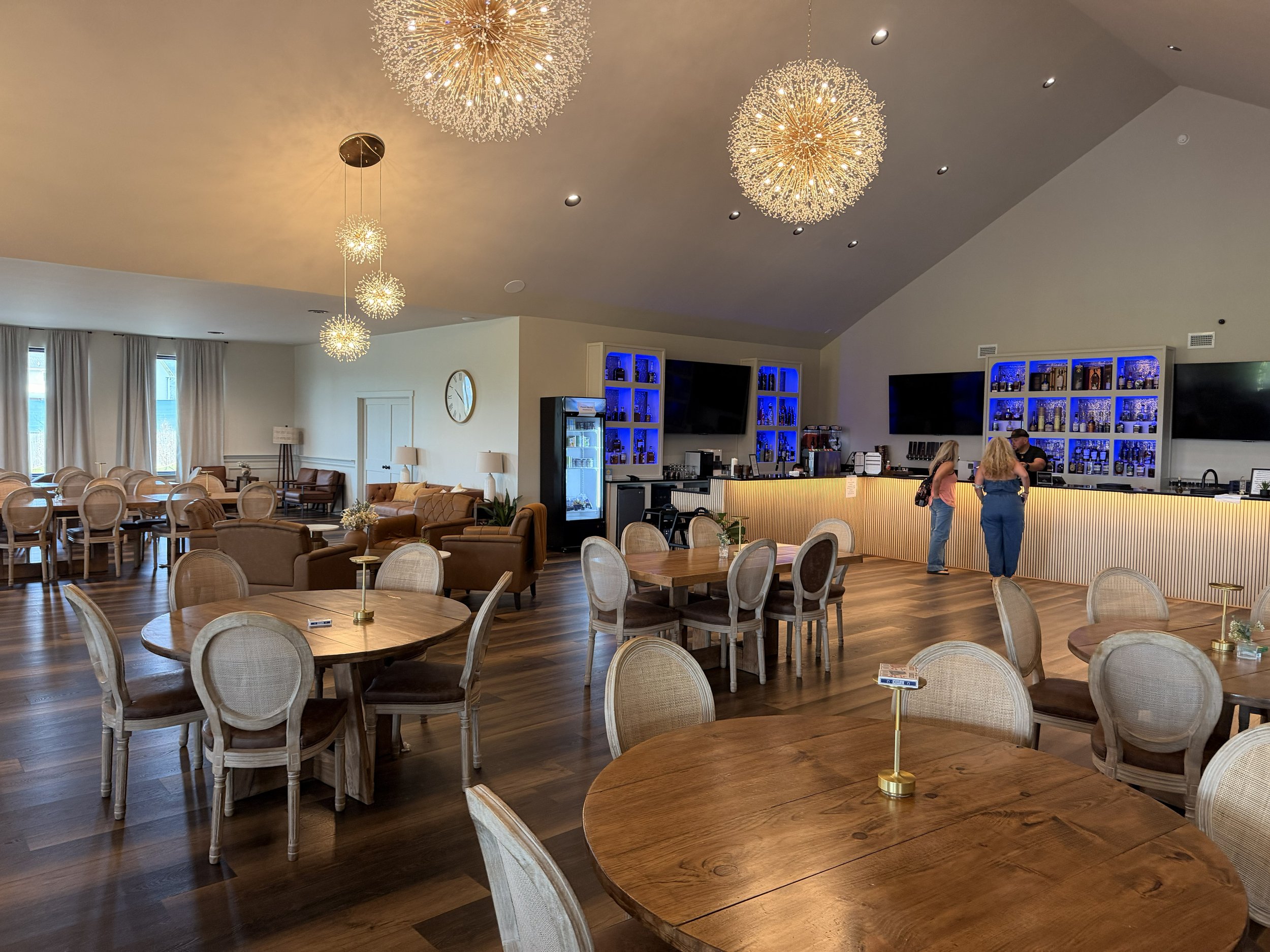 A spacious restaurant or cafe interior with round wooden tables and beige upholstered chairs. Modern chandeliers hanging from the ceiling illuminate the space, with some having sputnik-style designs and others with smaller spherical fixtures. A bar area with blue-lit shelves displaying bottles and glasses is visible on the right side, with two people standing at the bar counter. Large windows with curtains and a seating area with lounge chairs are in the background.