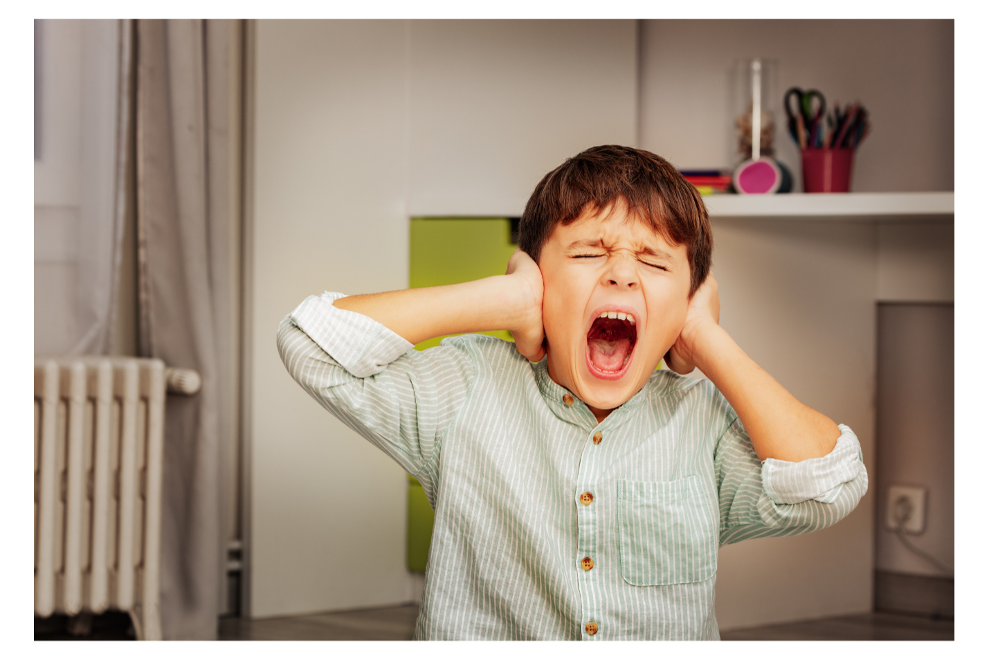 A young boy with brown hair, wearing a light green striped shirt, is covering his ears and yelling loudly in a room with shelves and a radiator.