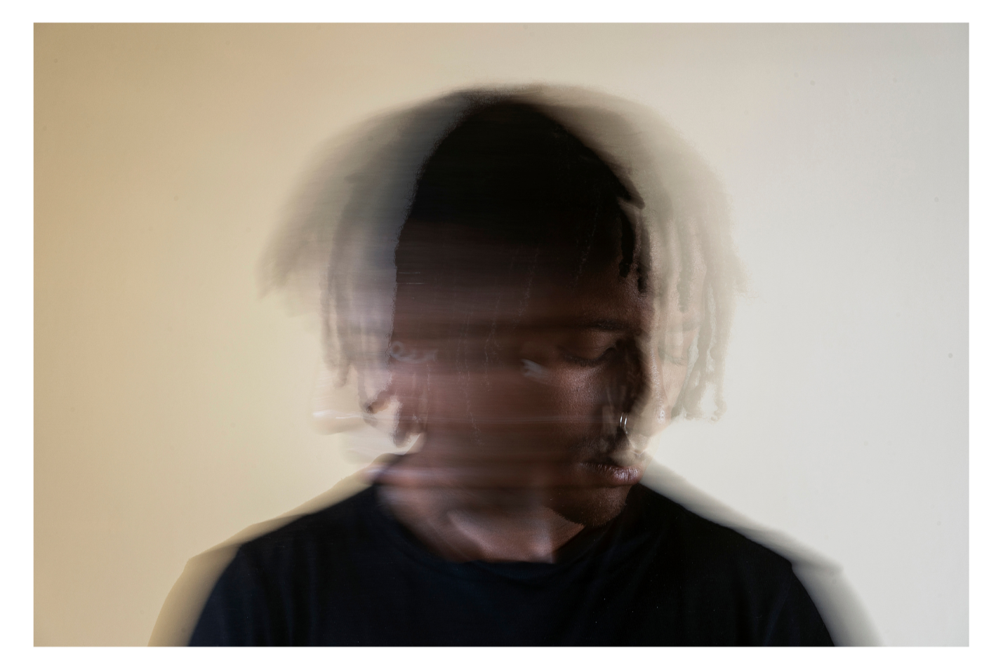 A person with dark skin and short dreadlocks wearing a black shirt, with another person's face superimposed in motion blur, creating a double exposure effect, against a plain light-colored background.