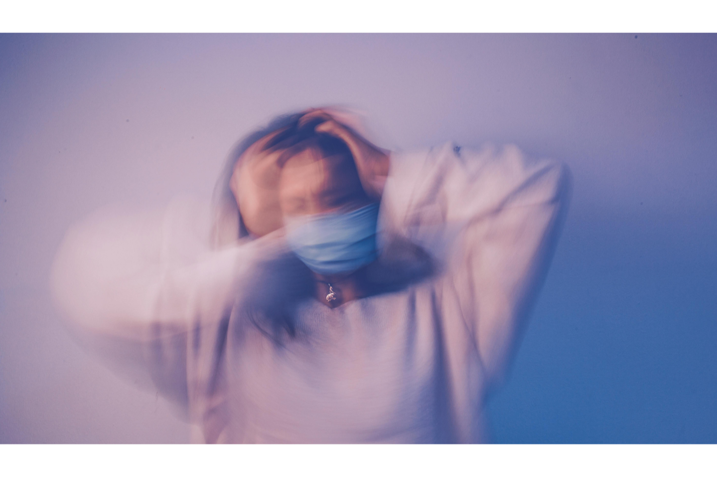 Person wearing a face mask holding their head with both hands, standing against a blue and purple background, with a blurry motion effect.