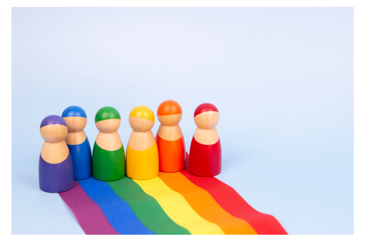 Six colorful wooden figurines arranged in a row on a light background, each casting a rainbow-colored shadow representing the LGBTQ+ pride flag.