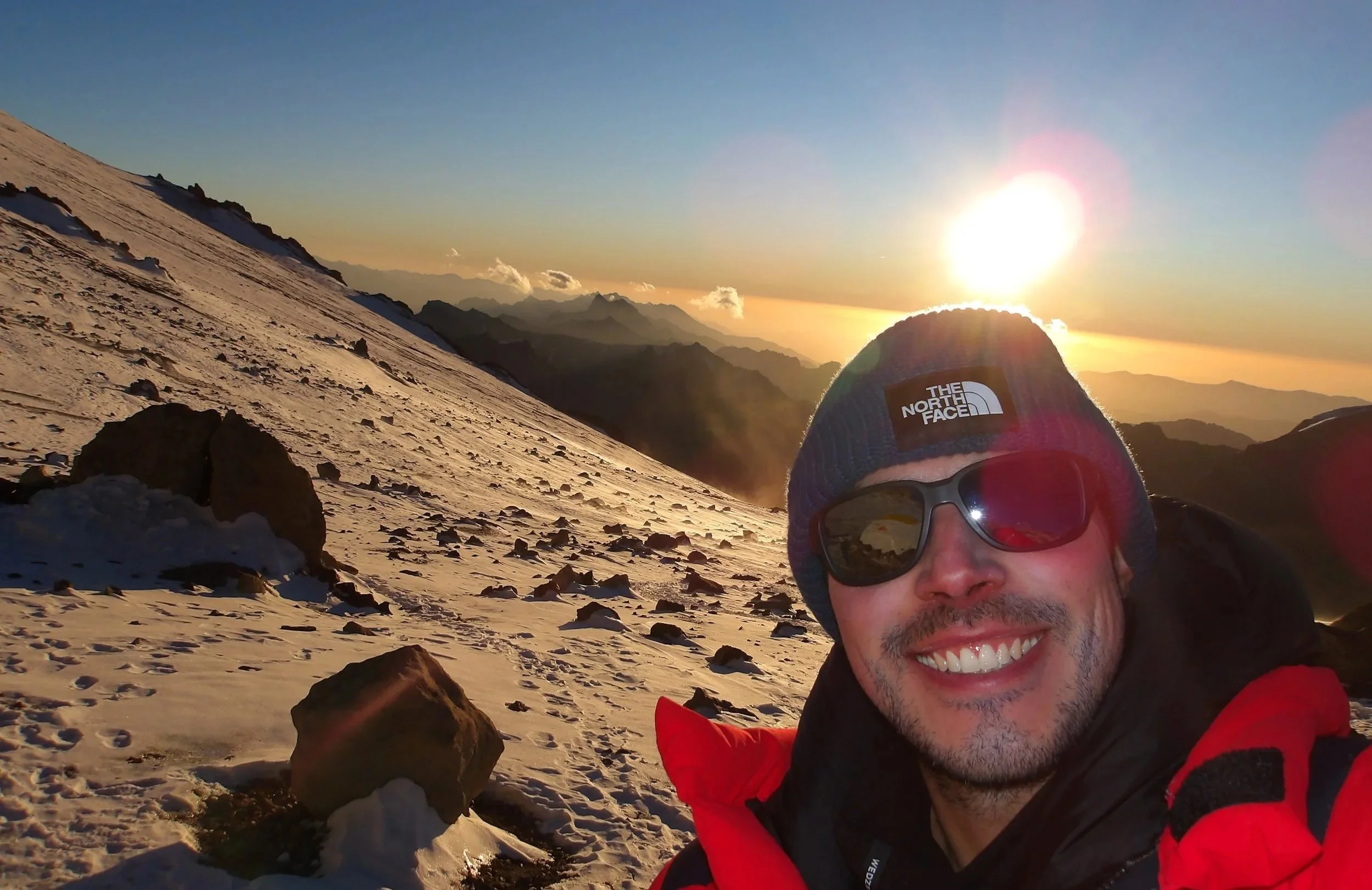 Hombre sonriendo con gafas y gorro en la cima de una montaña nevada al atardecer, con un paisaje montañoso en el fondo
