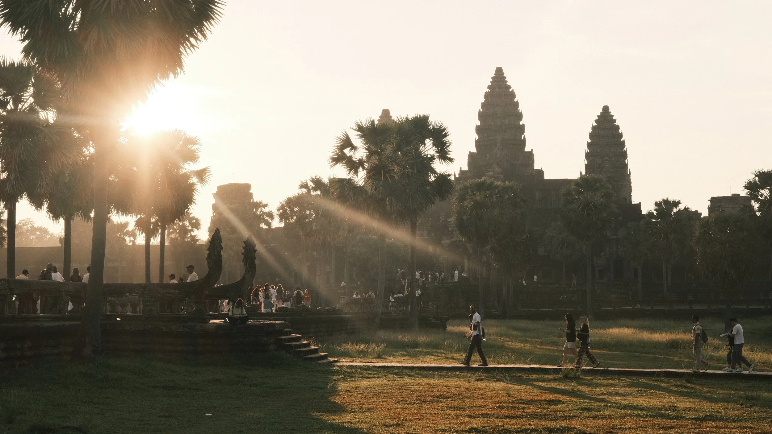 Templo de Angkor Wat al fondo, con persona caminando y los rayos del sol llegando entre palmeras