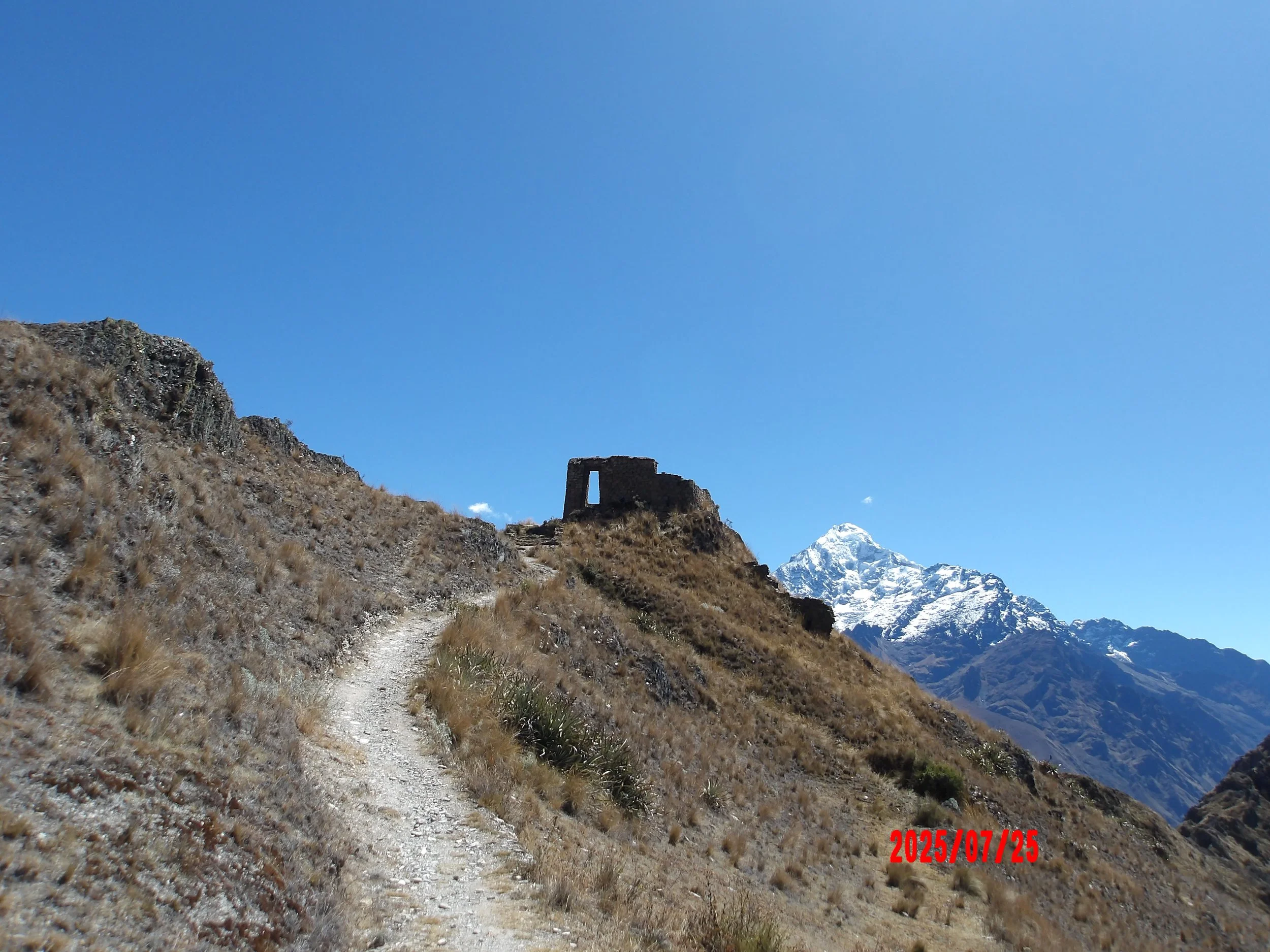 Sendero y ruinas de Inti Punku con montañas nevadas al fondo.