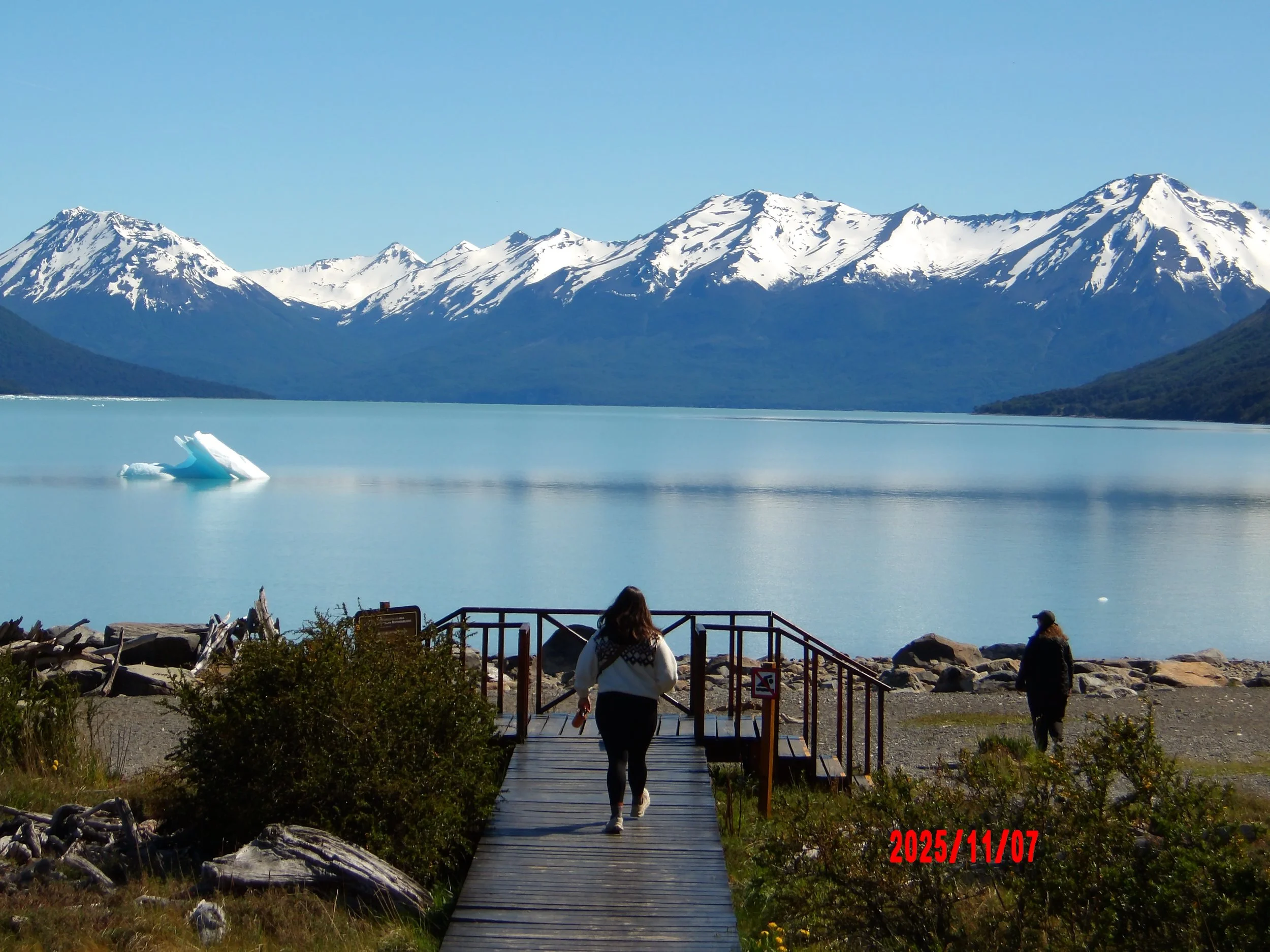 Mujer caminando sobre pasarela con el Lago Argentino al fondo y montañas nevadas.