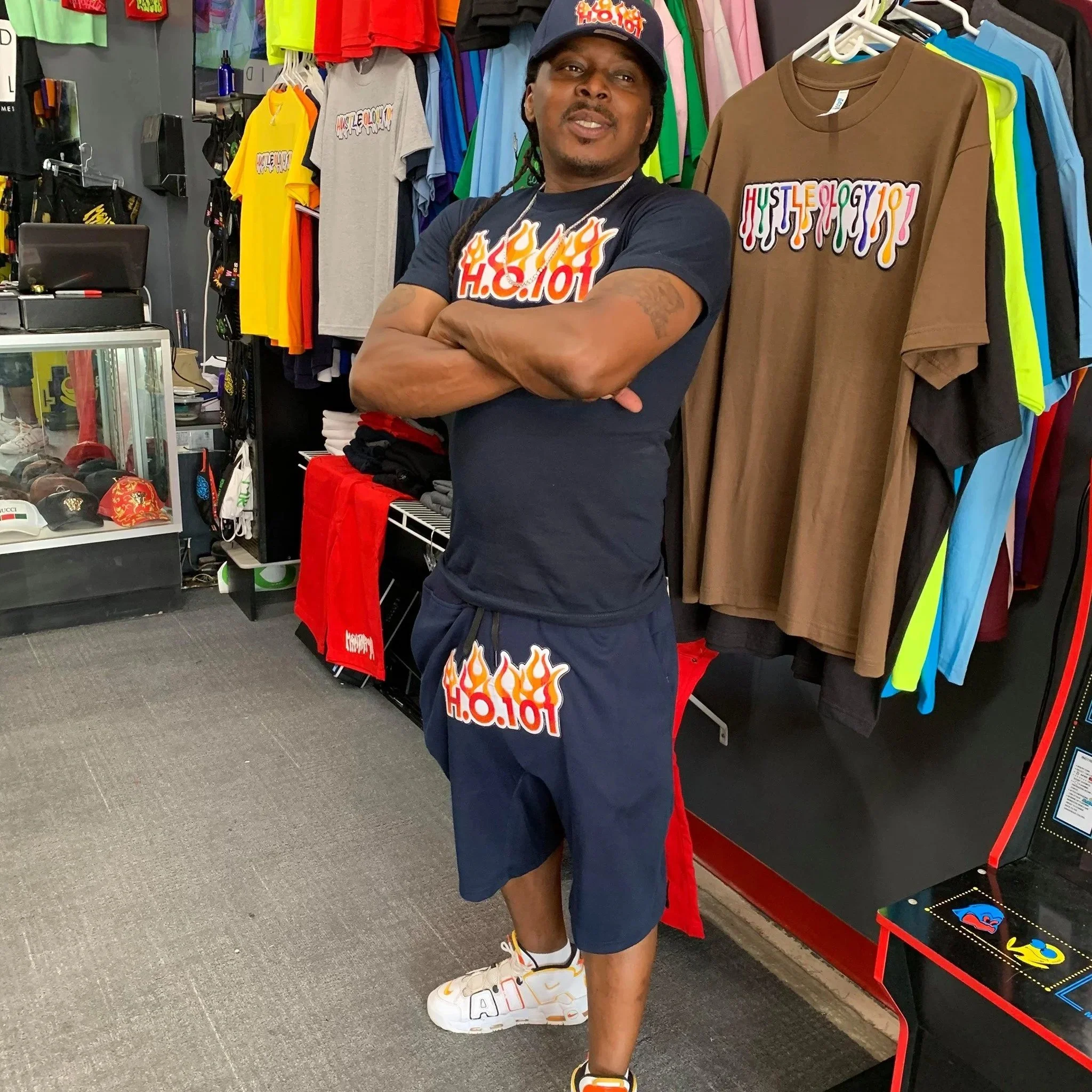 Man standing inside a clothing store, wearing a black t-shirt, navy shorts, white sneakers, and a black cap. The clothing store has brightly colored t-shirts and sweatshirts hanging on racks behind him and a display case with hats to his left.