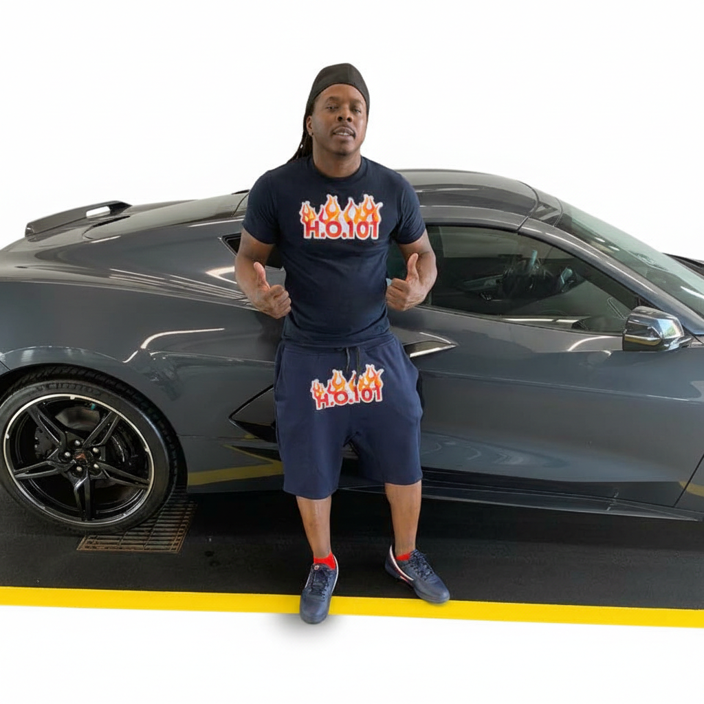Man standing in front of a gray sports car, wearing a black head covering, navy blue t-shirt and shorts with 'H.O.T' flame logo, giving a thumbs up.