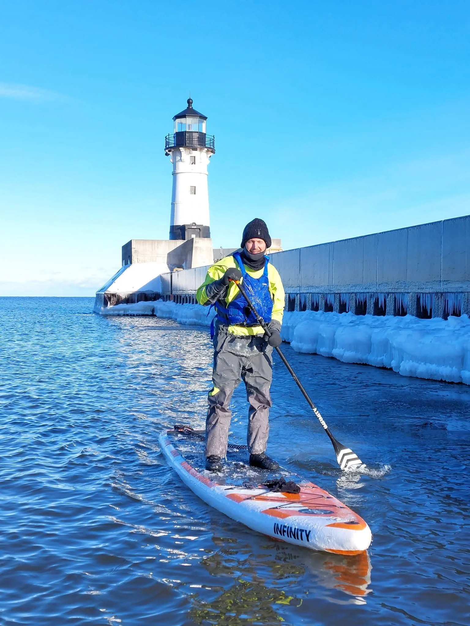 Person in winter gear paddleboarding near a lighthouse on icy waters.