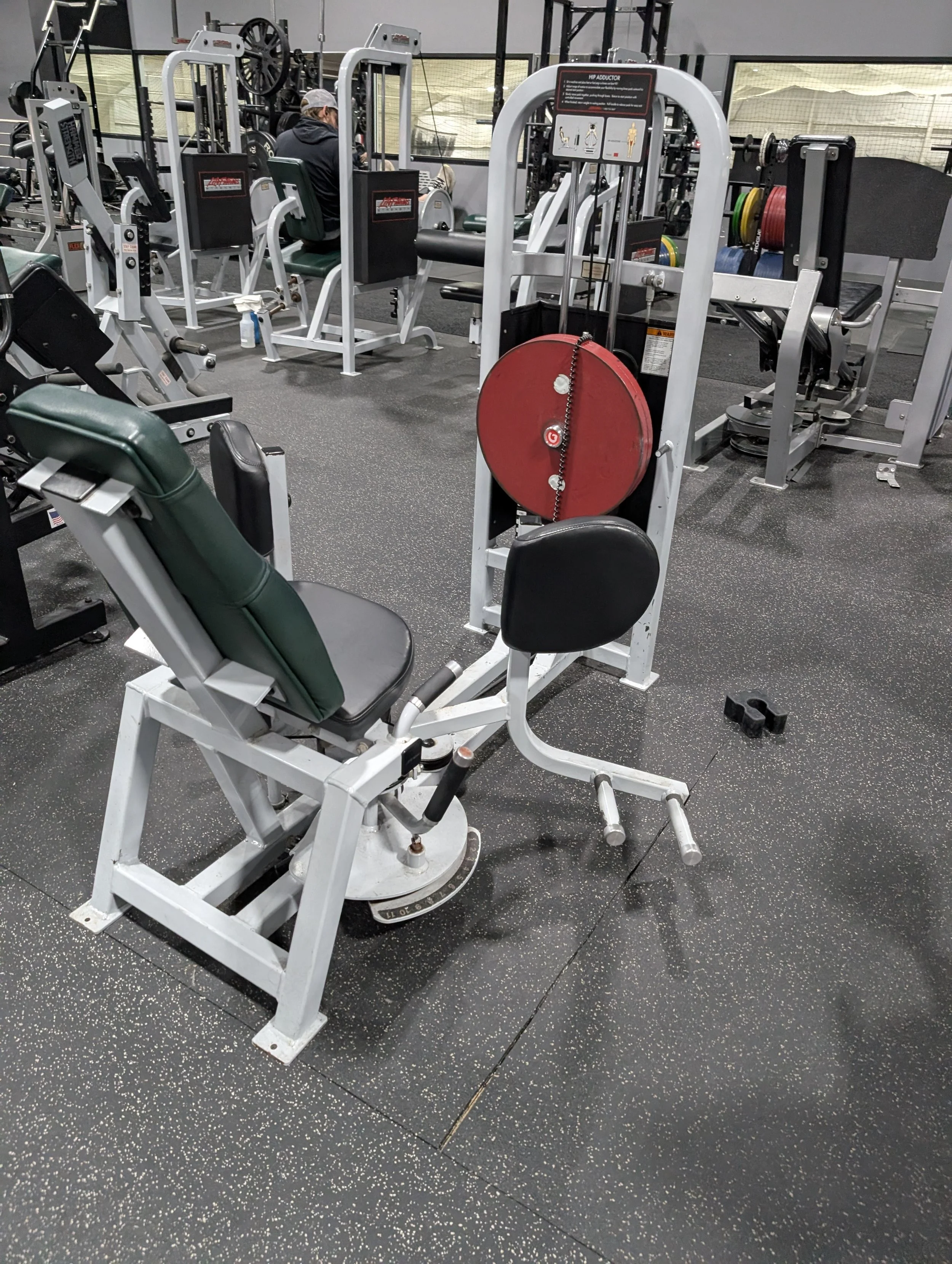 An old-fashioned strength training machine with a red weight plate, black padded supports, and a chair for seated exercise, inside a gym with other equipment and a person working out in the background.