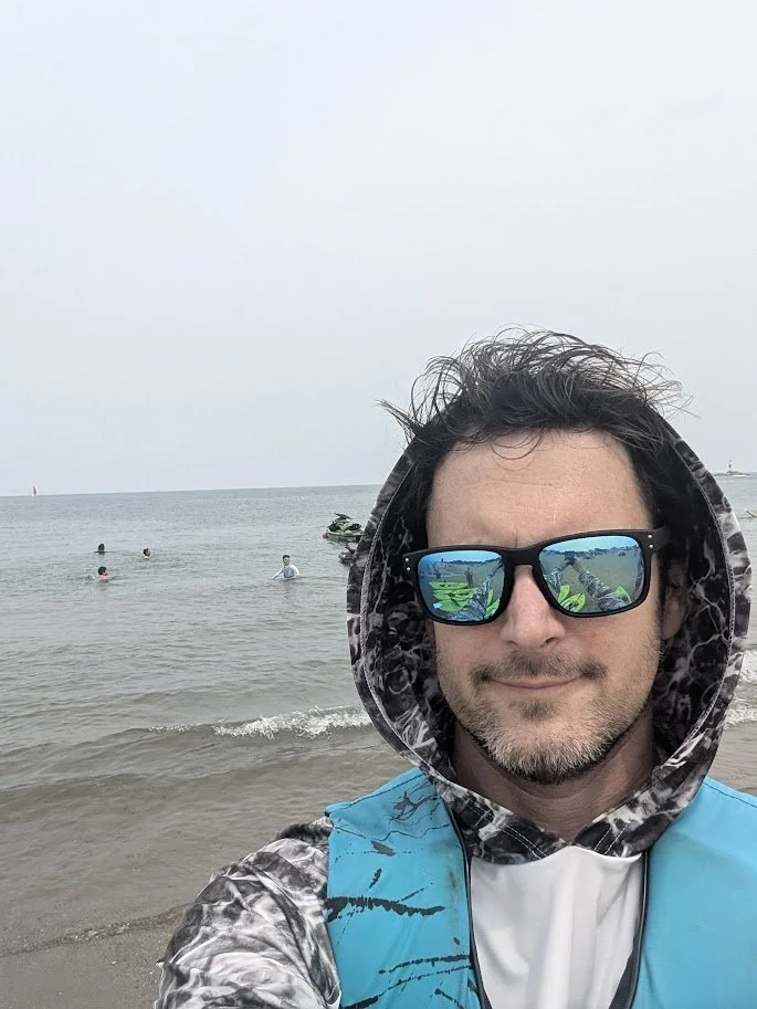 A man taking a selfie on the beach, wearing sunglasses, a hoodie, and a life vest, with the ocean and a few people swimming in the background.