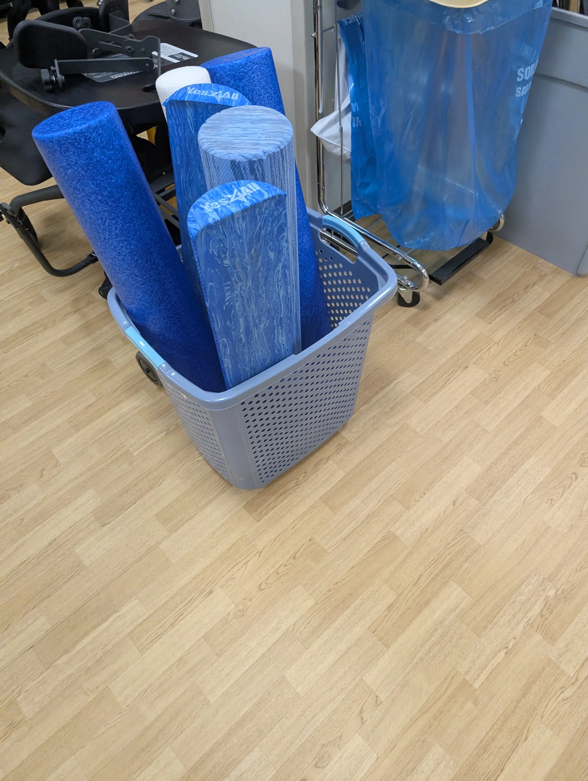Blue cleaning mop handles stacked in a grey plastic basket on a wooden floor next to a blue trash bag.