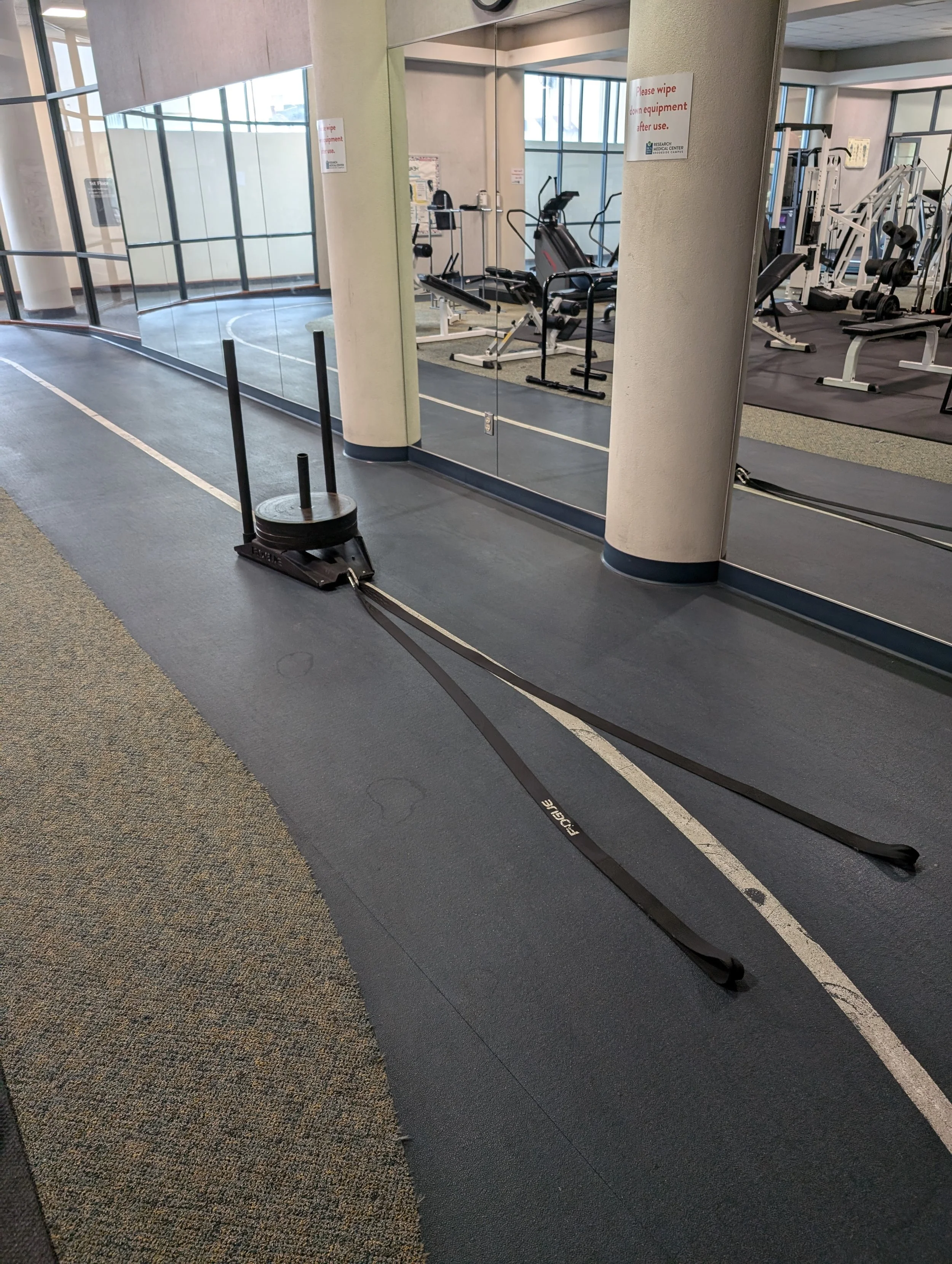 A fitness center with a sled and resistance bands on the gym floor near a glass wall with exercise equipment visible in the background.