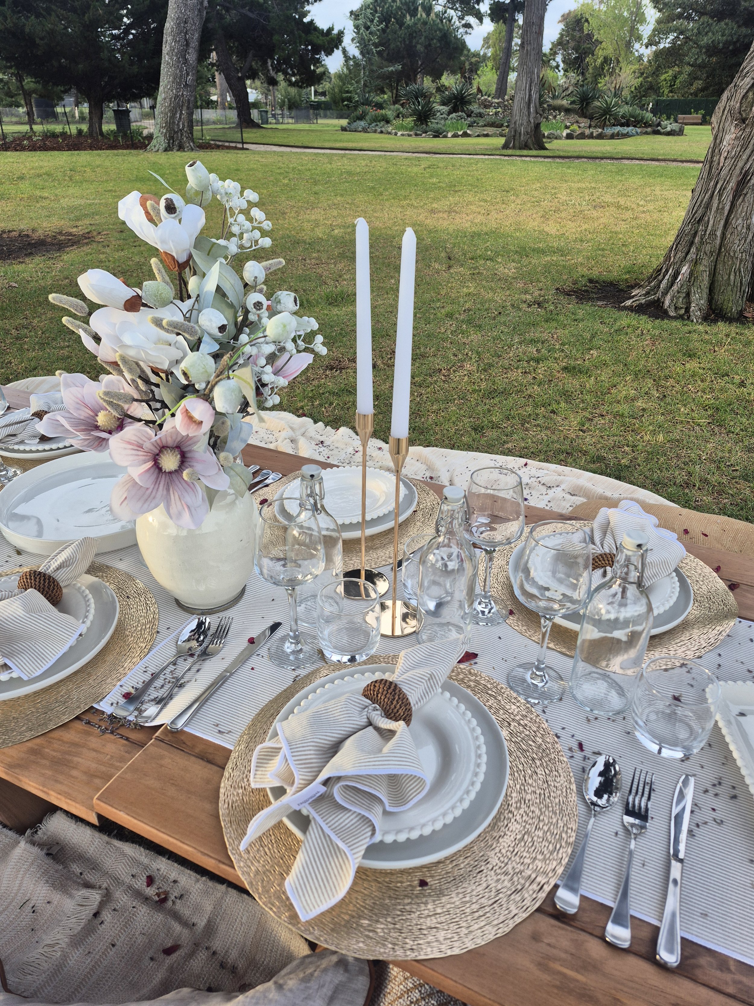 Elegant outdoor picnic setting featuring premium tableware, decorative florals, candles, and beautifully styled details surrounded by natural parkland.