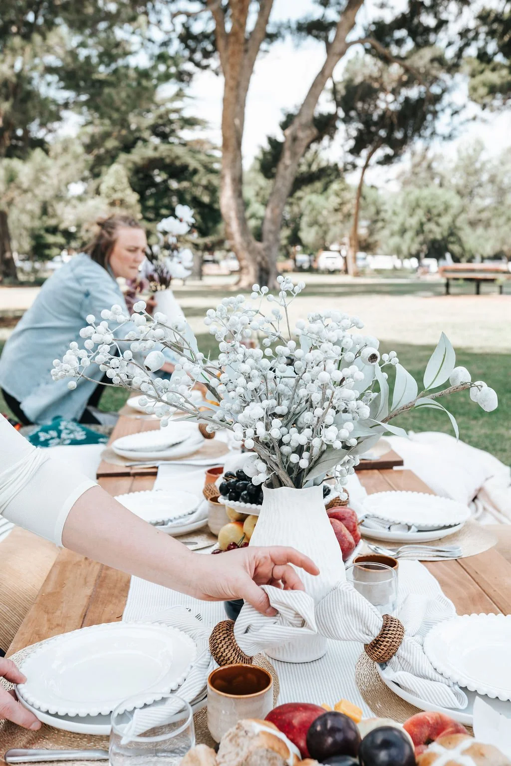 Elegant picnic table styling featuring fresh seasonal fruit, premium tableware, decorative florals, and beautifully considered details in a scenic outdoor setting.