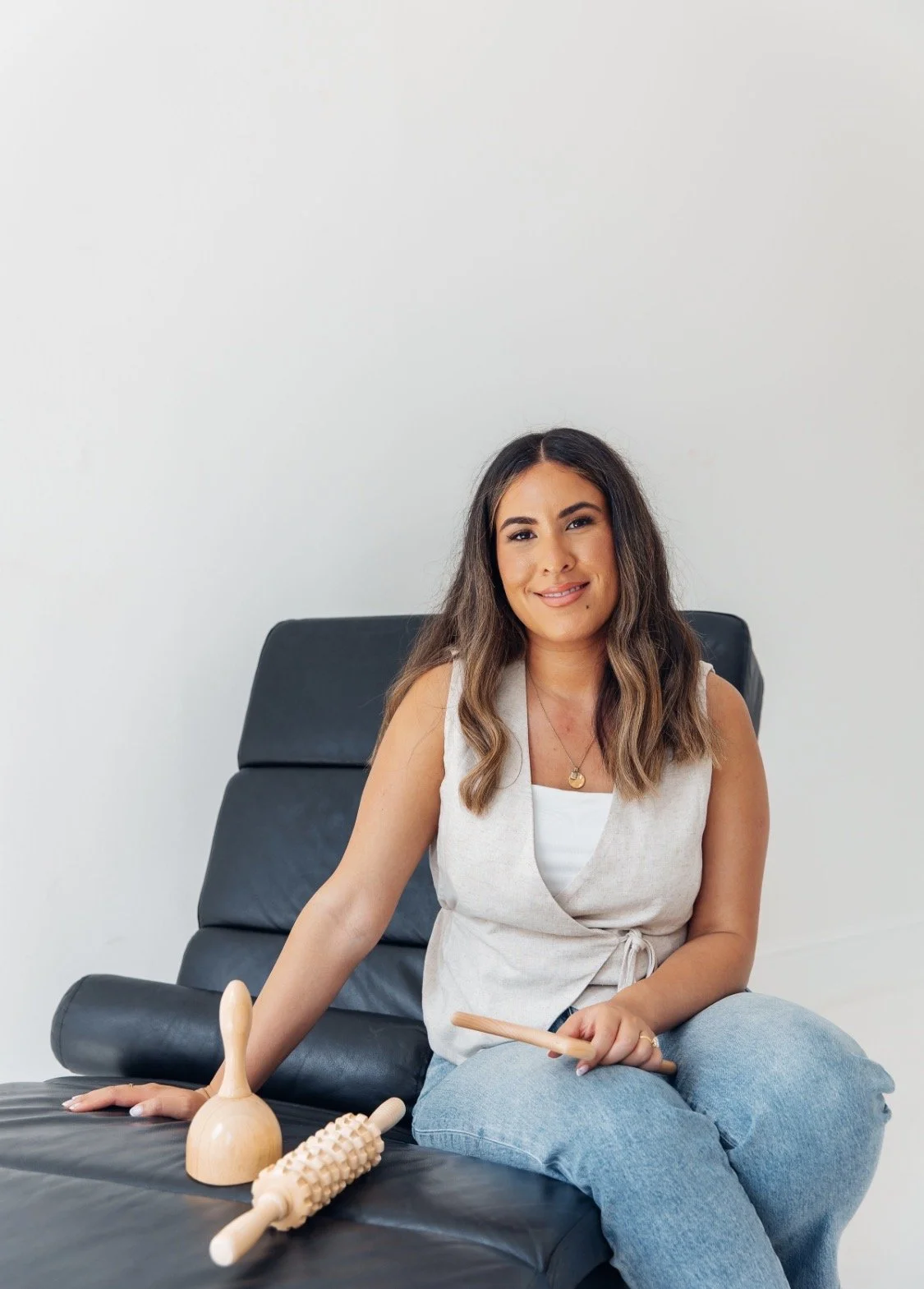 A woman sitting on a black leather massage chair holding a wooden massage tool, with other wooden massage tools placed on the chair in front of her, against a plain white wall.