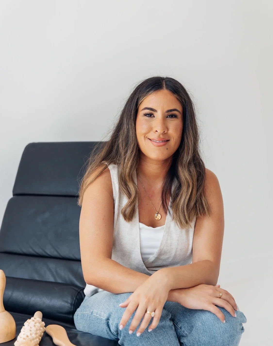 A woman with shoulder-length brown hair, wearing a sleeveless white top and blue jeans, seated on a black massage chair with skincare tools nearby, smiling at the camera.
