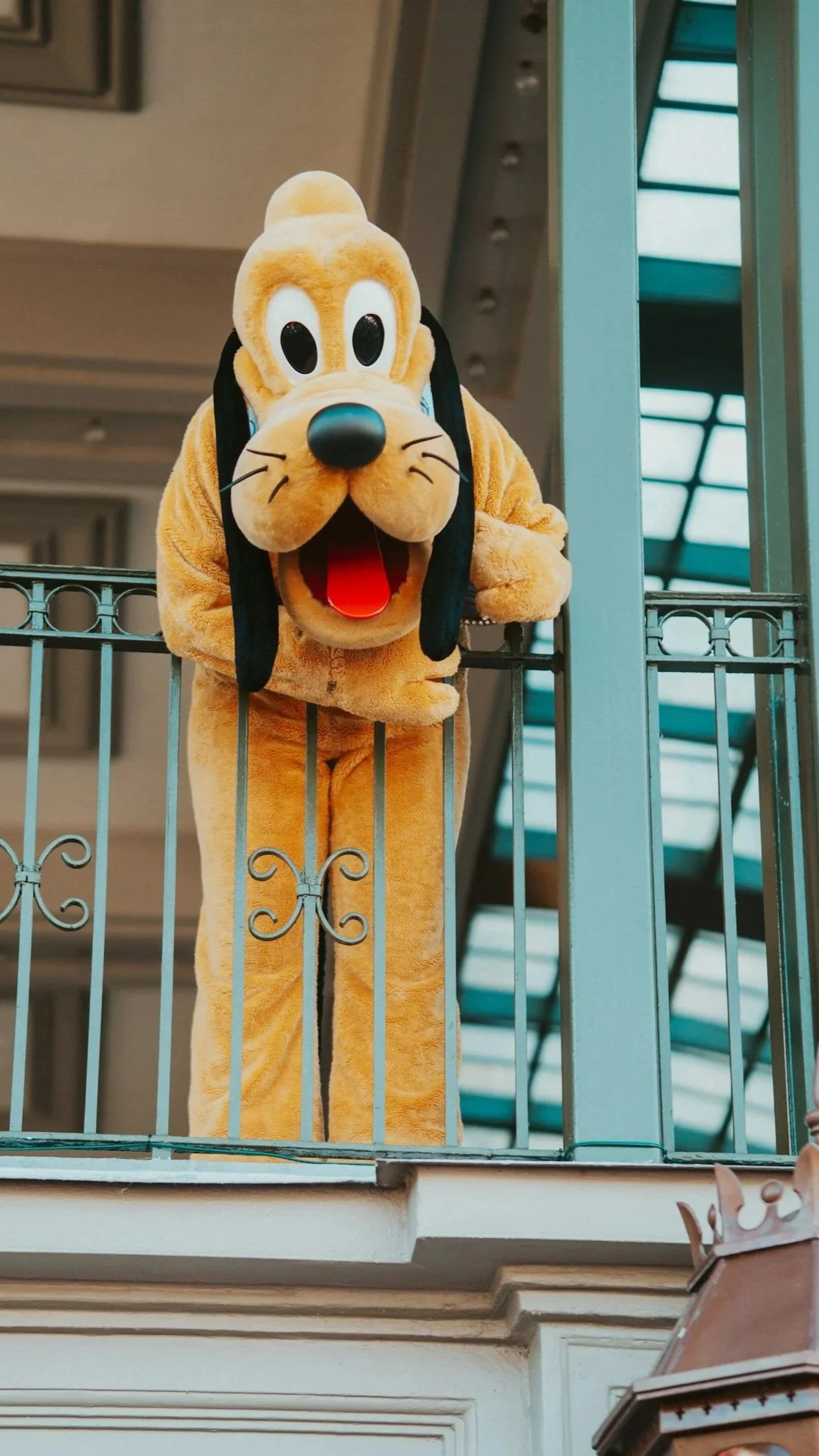 Costumed mascot of Pluto, the Disney character, leaning over a balcony with a playful expression. The mascot is in a tan, furry costume with floppy black ears, a large black nose, and a red tongue visible in an open-mouthed smile.