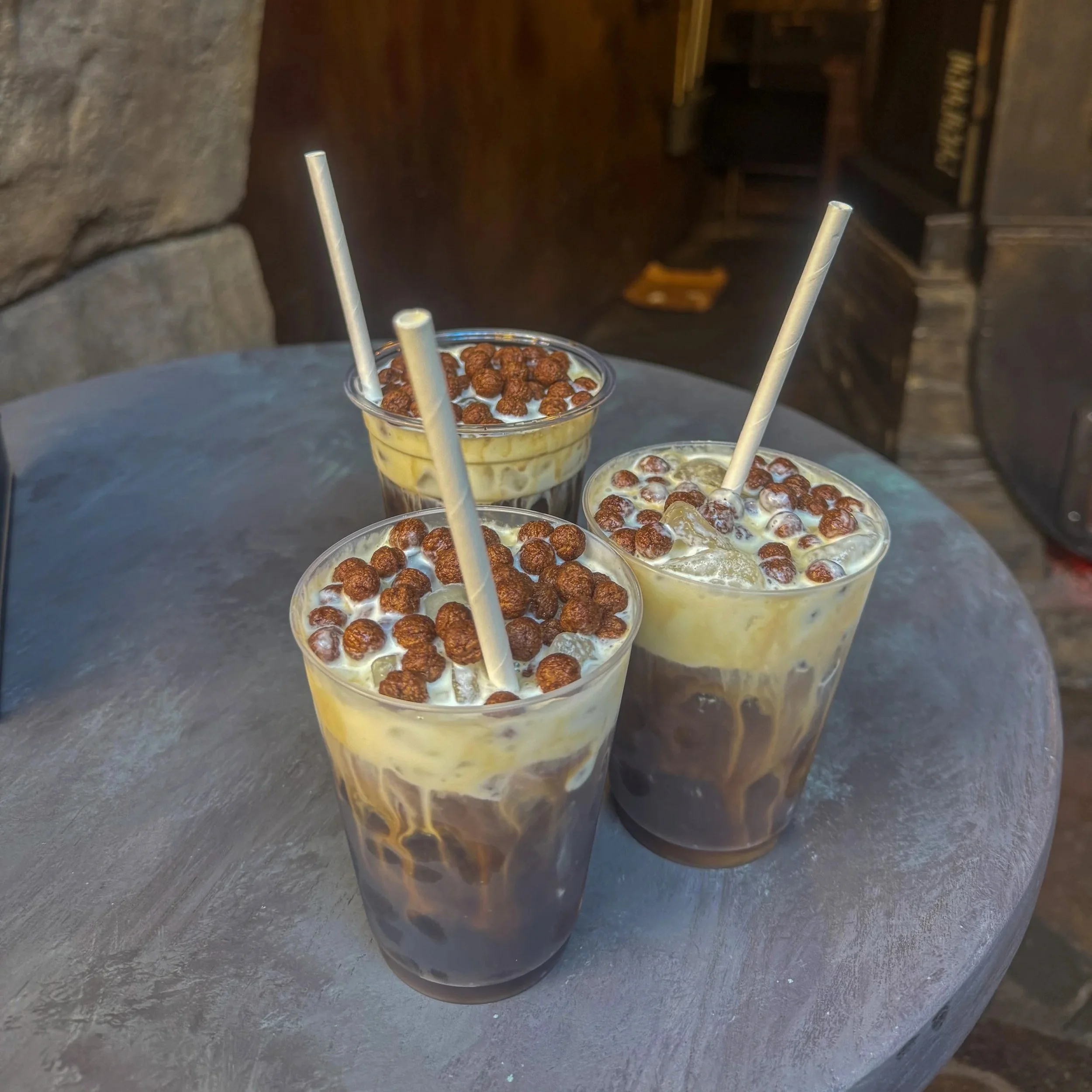 Three glasses of iced coffee topped with chocolate cereal balls on a round table.