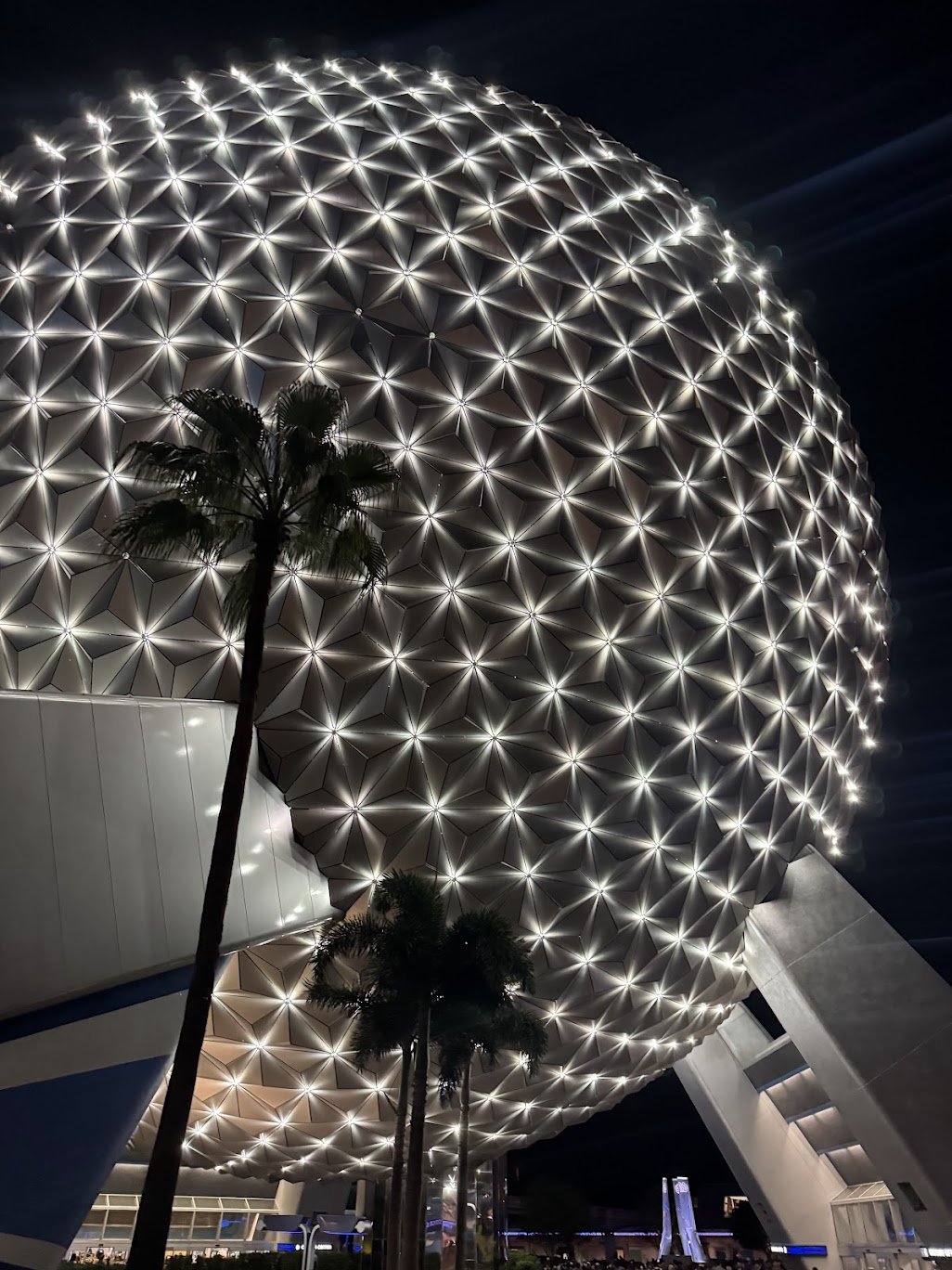 Nighttime view of a large geodesic sphere with illuminated triangular panels, topped with a pattern of bright lights, with palm trees in the foreground.