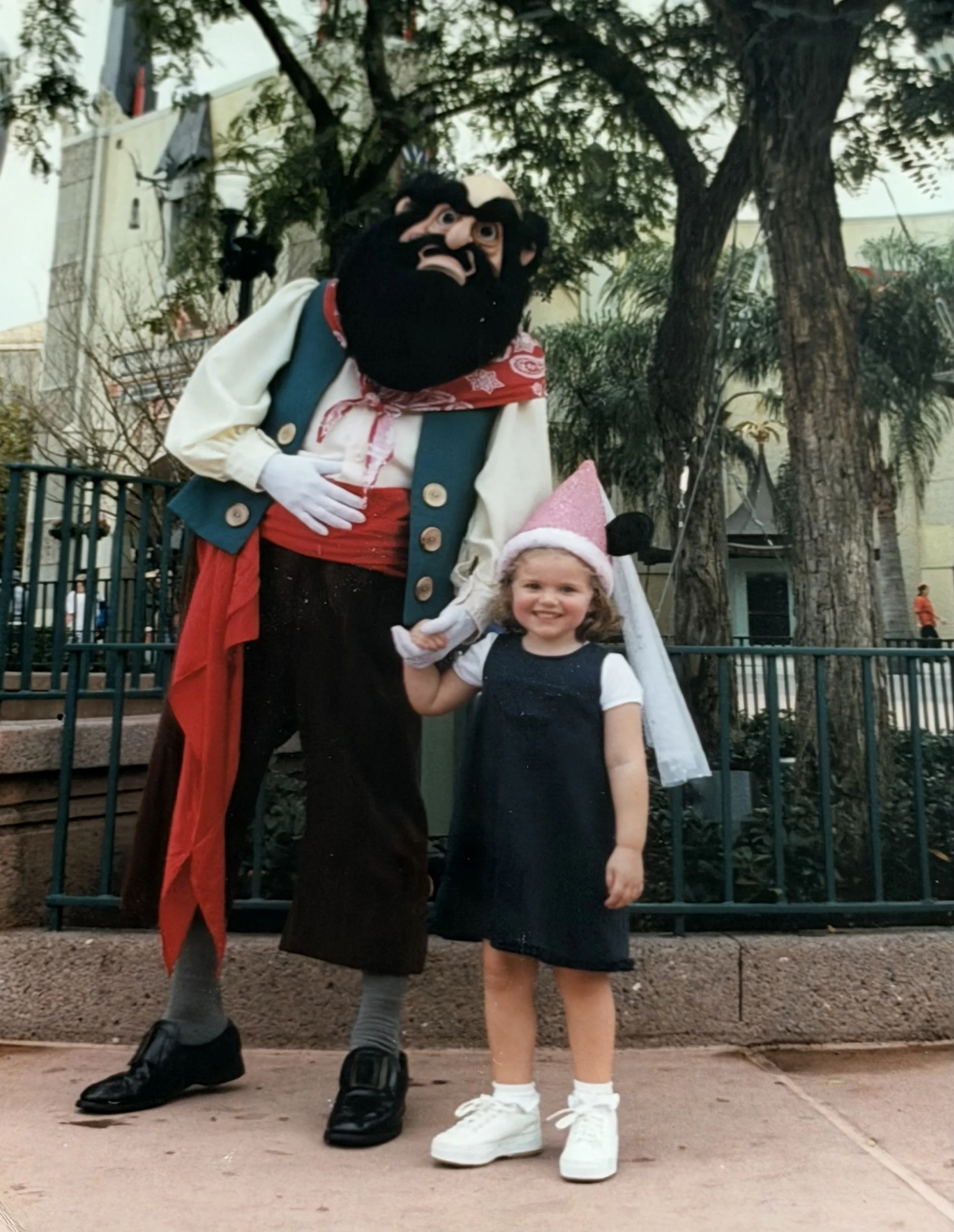 Young girl smiling and holding hands with a costumed character of a pirate with a large beard and bandana, in an outdoor park setting.