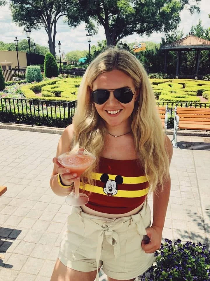 A young woman with long blonde hair, wearing sunglasses, a Mickey Mouse shirt, and beige shorts, holding a pink cocktail glass, standing outdoors in a park-like setting with greenery, trees, benches, and a gazebo.