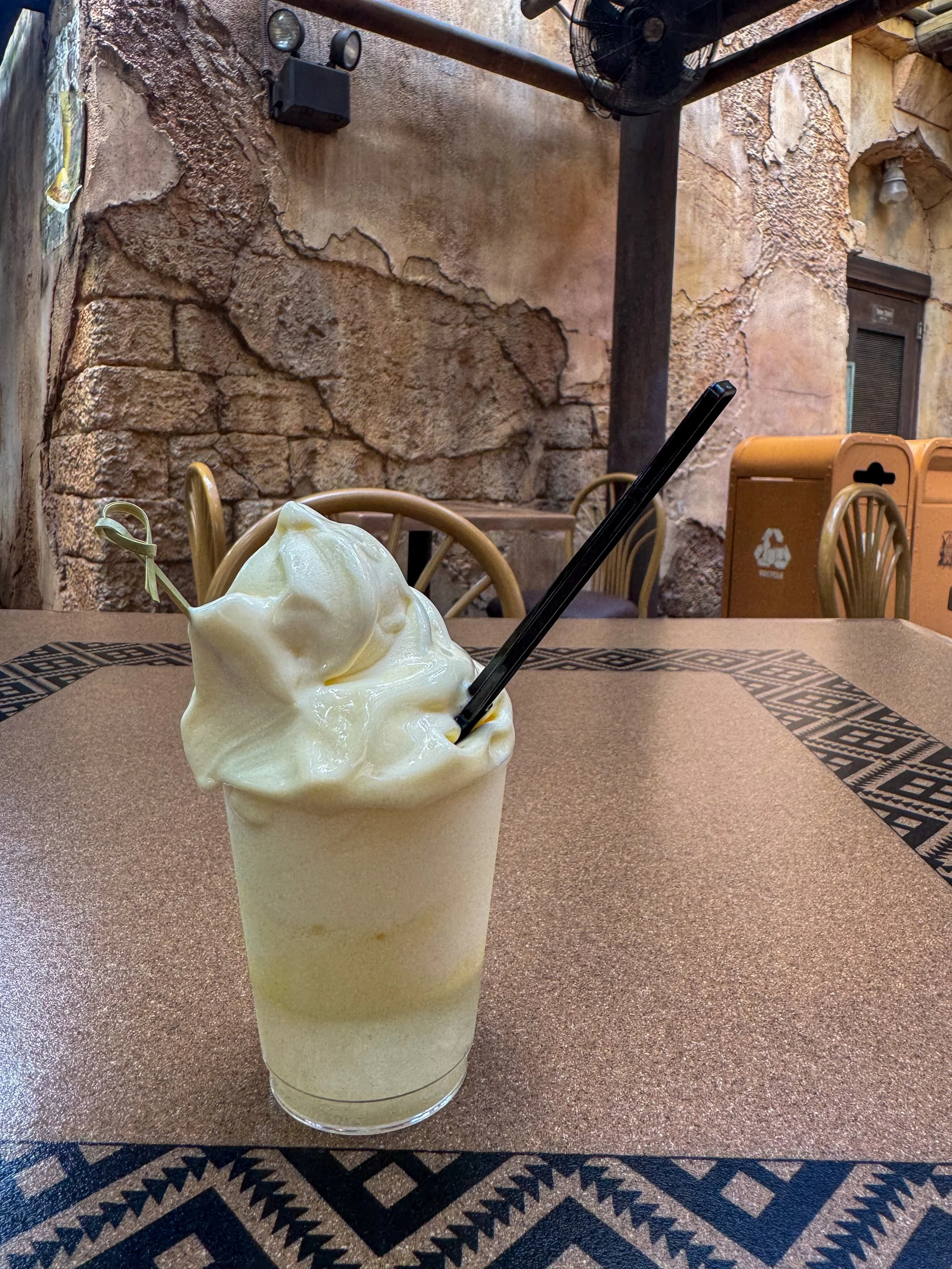A creamy, pale yellow smoothie with soft serve ice cream on top and a black straw, served in a clear glass on a patterned table in a rustic outdoor café setting.