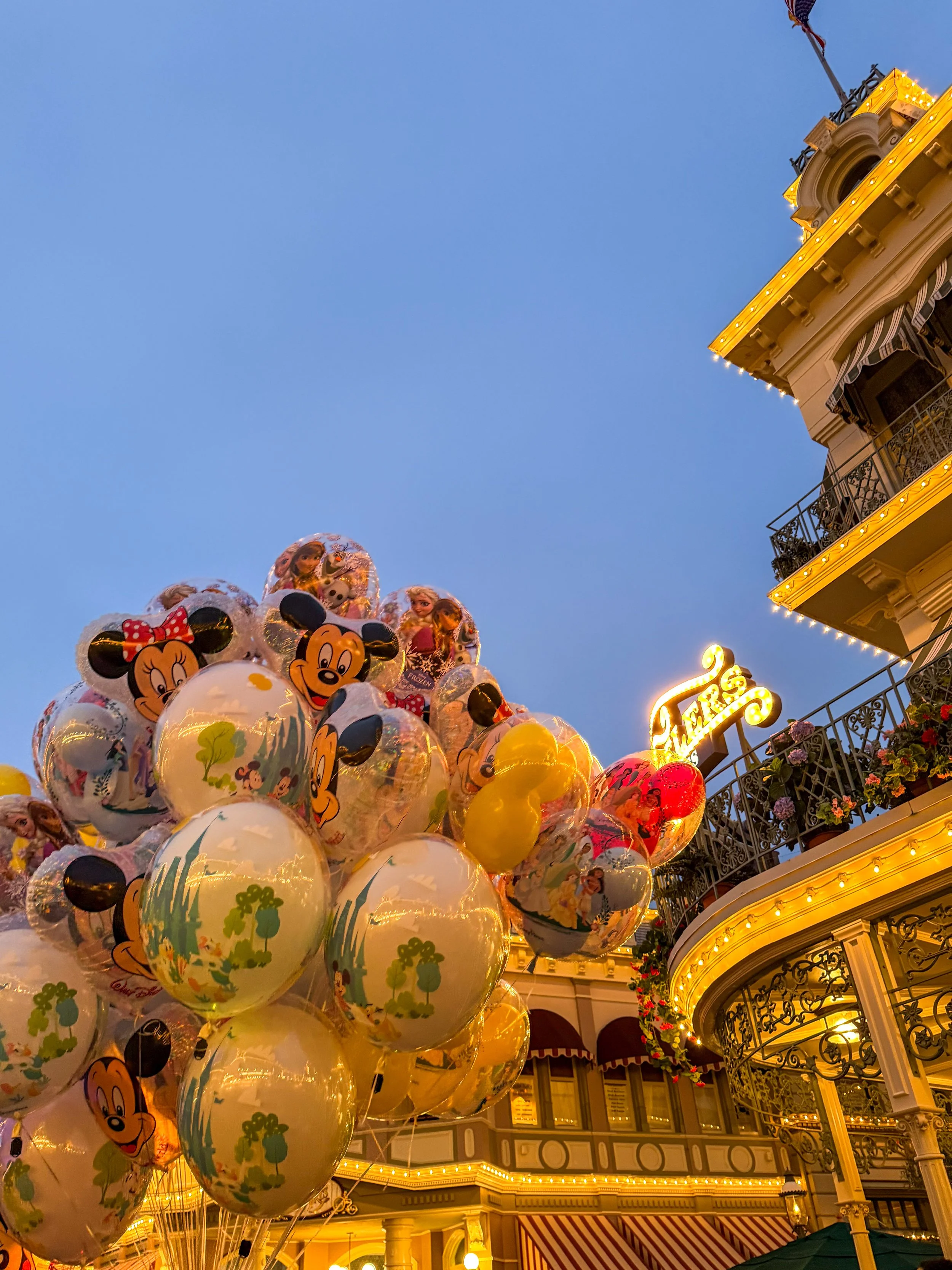 Balloon display featuring Disney characters like Mickey Mouse and Minnie Mouse at Disneyland during twilight, with a decorative building adorned with lights in the background.