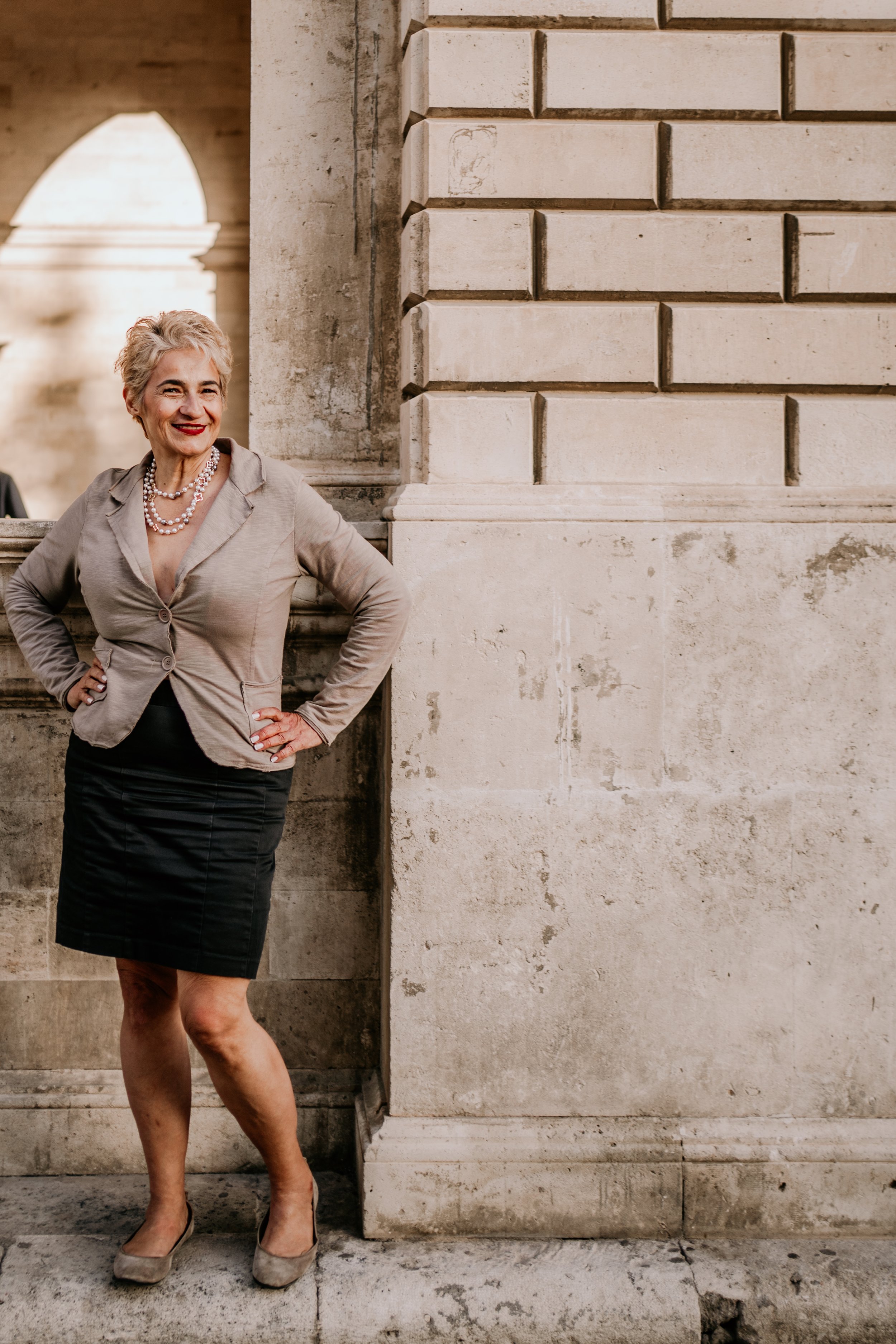 A woman with short gray hair and red lipstick, wearing a beige blazer, black skirt, and beige heels, stands with her hands on her hips in front of a stone wall with classical architectural features.