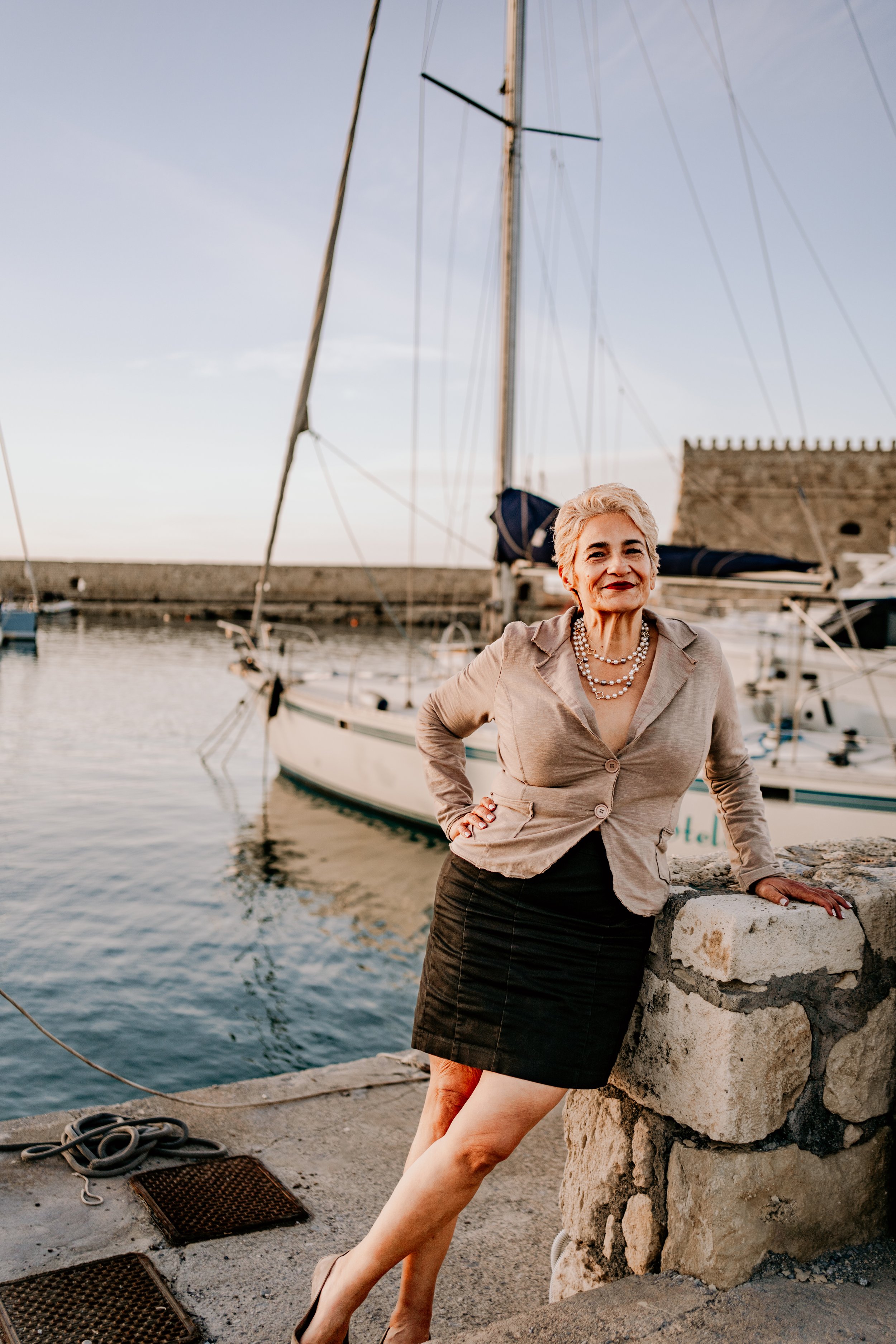A woman with short blonde hair wearing a beige blazer and black skirt, standing on a stone pier by the water, smiling, with sailboats and a stone building in the background during sunset.